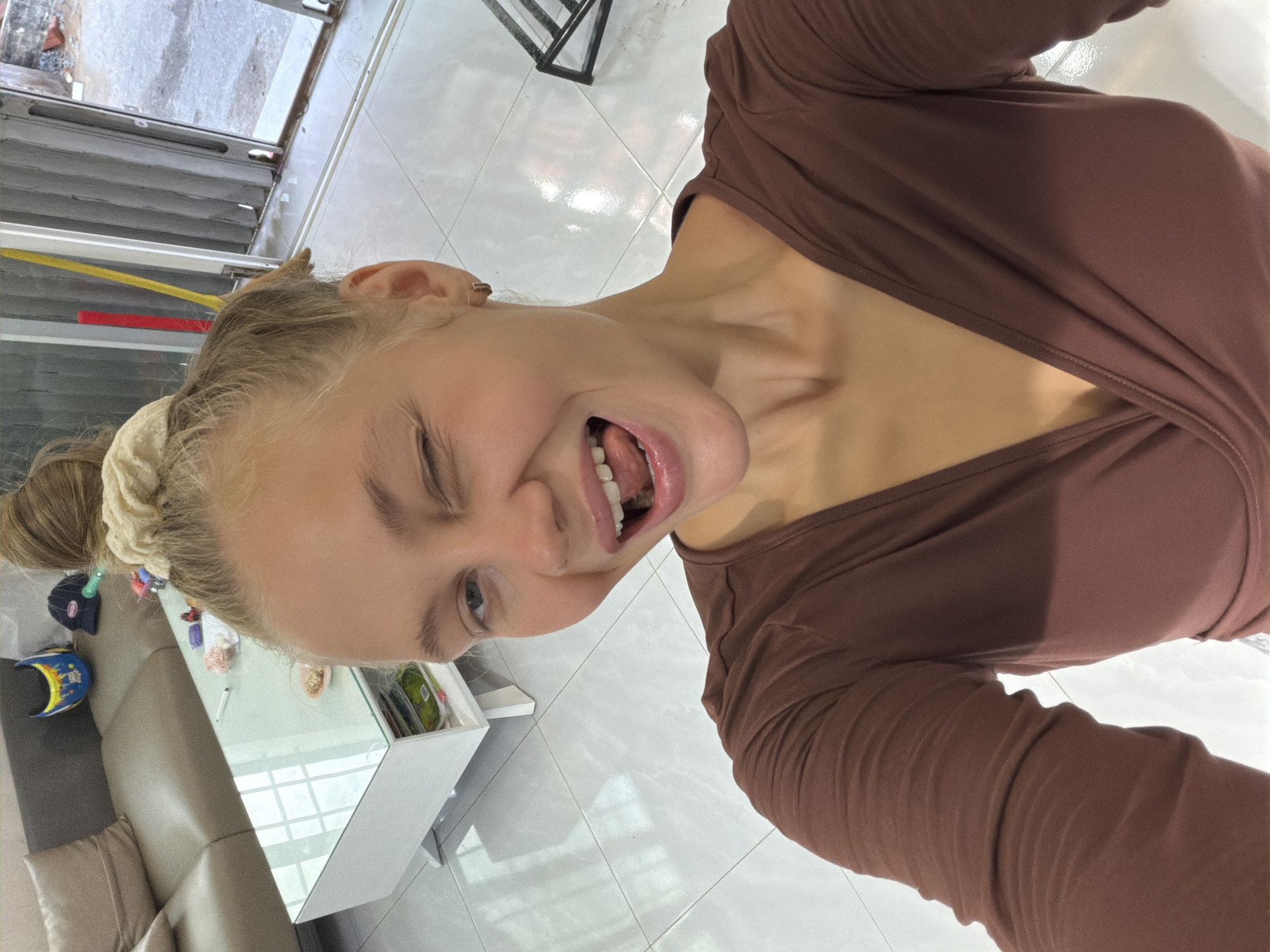 A young Caucasian woman with blonde hair pulled up in a messy bun is taking a selfie in what appears to be a modern living room. She is winking and sticking her tongue out, wearing a brown long-sleeved, low-cut wrap top. The room features a large, light-colored sectional sofa, a white coffee table with a glass top, and a tiled floor. In the background, through an open doorway or window, there is an outdoor view of a street and buildings, suggestive of an urban setting. The presence of a couch and coffee table indicates an indoor, residential environment. The overall impression is a casual, playful moment captured in a domestic space.