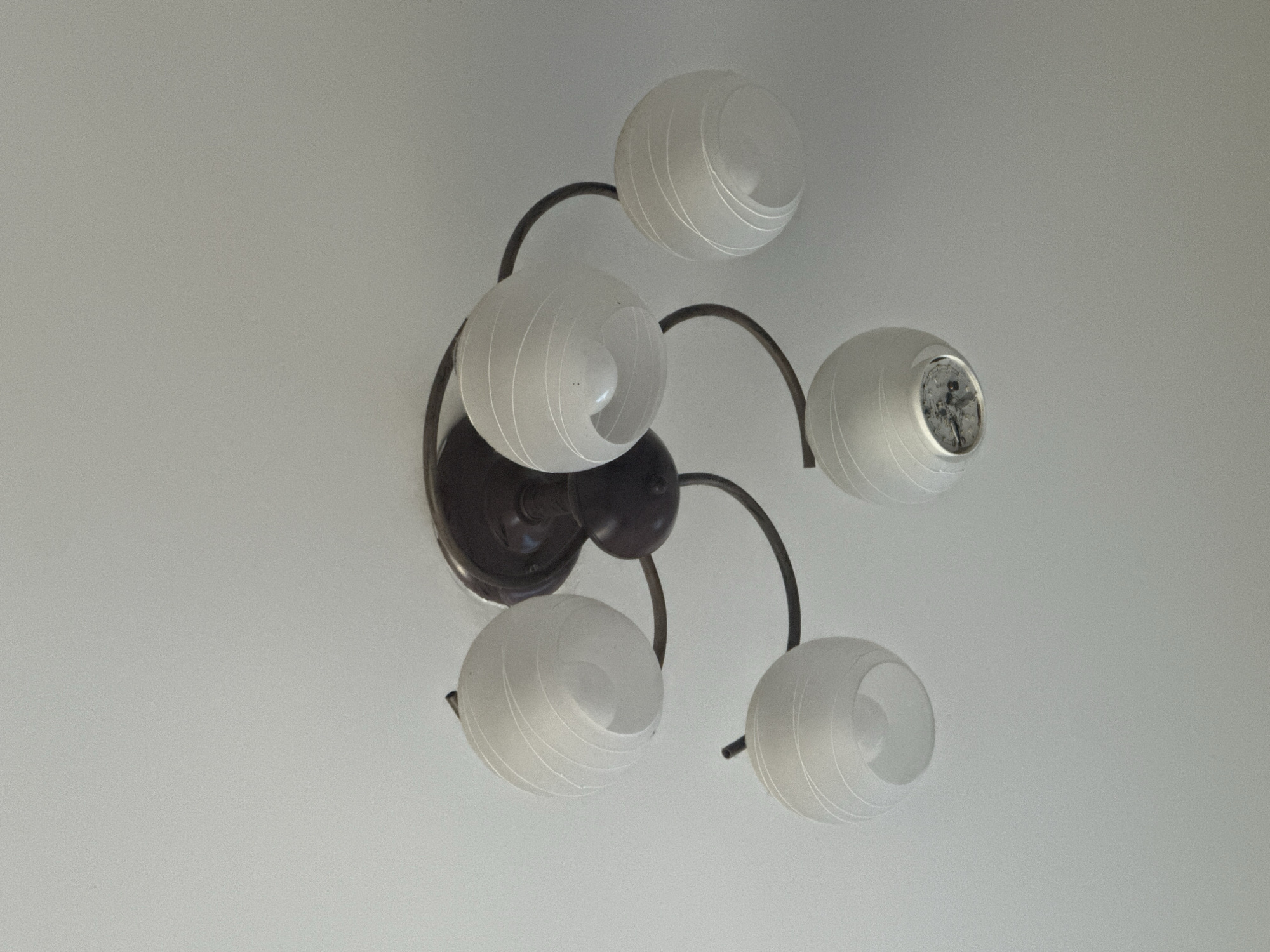 A ceiling light fixture hangs from a white ceiling. The fixture has a dark brown, circular base from which four dark metal arms extend. Each arm curves and supports a frosted white glass globe, which has a ribbed texture. Two of the globes appear to have light bulbs inside, one of which is lit. Another globe has a dark object visible inside, possibly a damaged bulb or fixture component. The overall design of the light fixture is ornate and somewhat vintage. The scene is indoors, and the lighting suggests it could be daytime, with the natural light supplementing the artificial light from the fixture. The setting appears to be a domestic interior, possibly a living room or hallway, in Karakol City, Kyrgyzstan. There is no visible text in the image.
