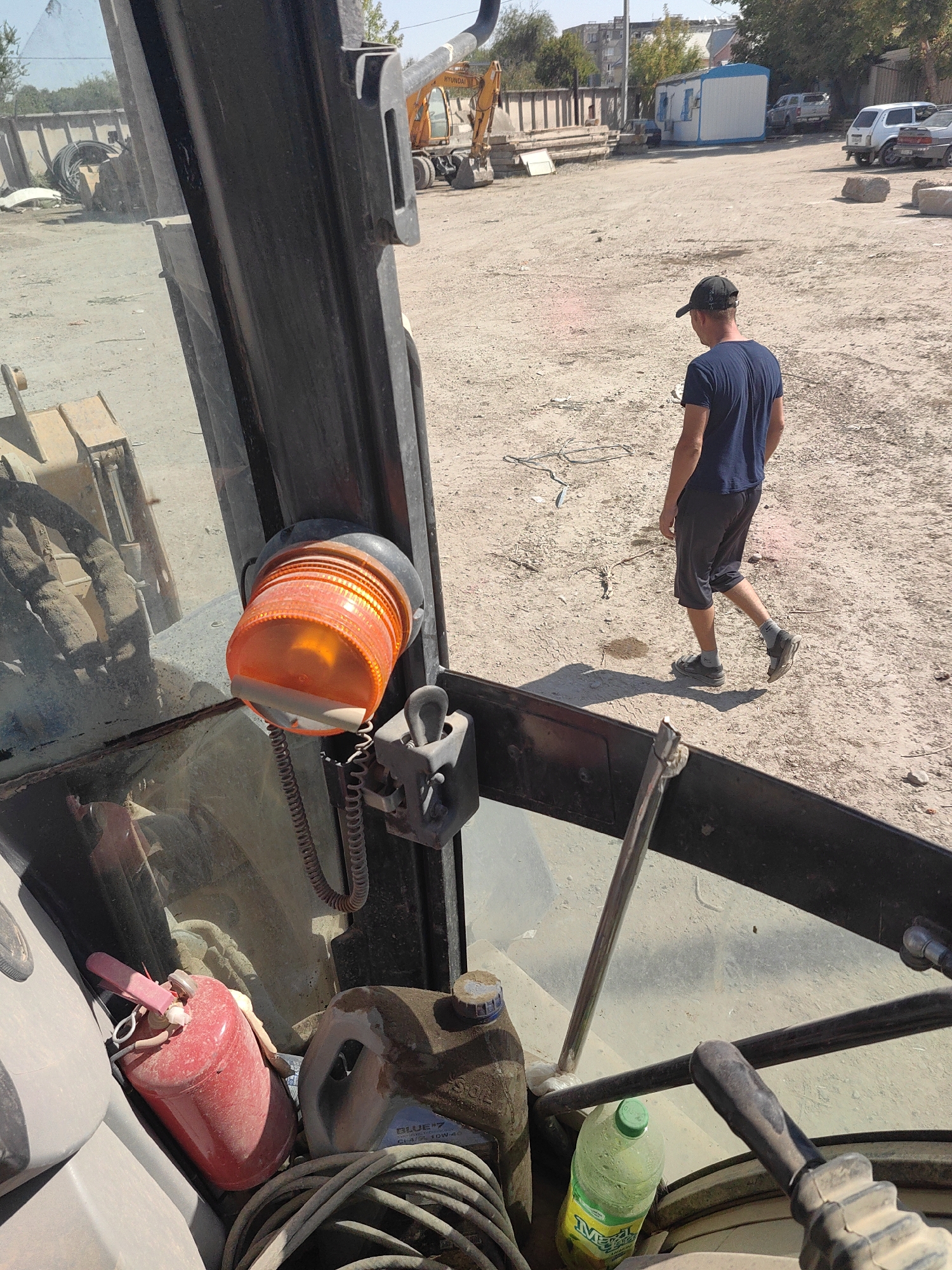A man wearing a dark blue t-shirt and dark shorts walks away from the camera in an outdoor, dusty environment that appears to be a construction or storage yard. He is wearing a dark baseball cap and grey socks with dark shoes. In the foreground, the interior of a vehicle, likely a construction machine, is visible, including an orange warning light, a red canister, and a green plastic bottle. In the background, a yellow excavator, several parked cars, a small portable building, and stacks of materials are present. The scene is illuminated by natural daylight under a clear sky. The location is specified as a construction yard in Shymkent, Kazakhstan.