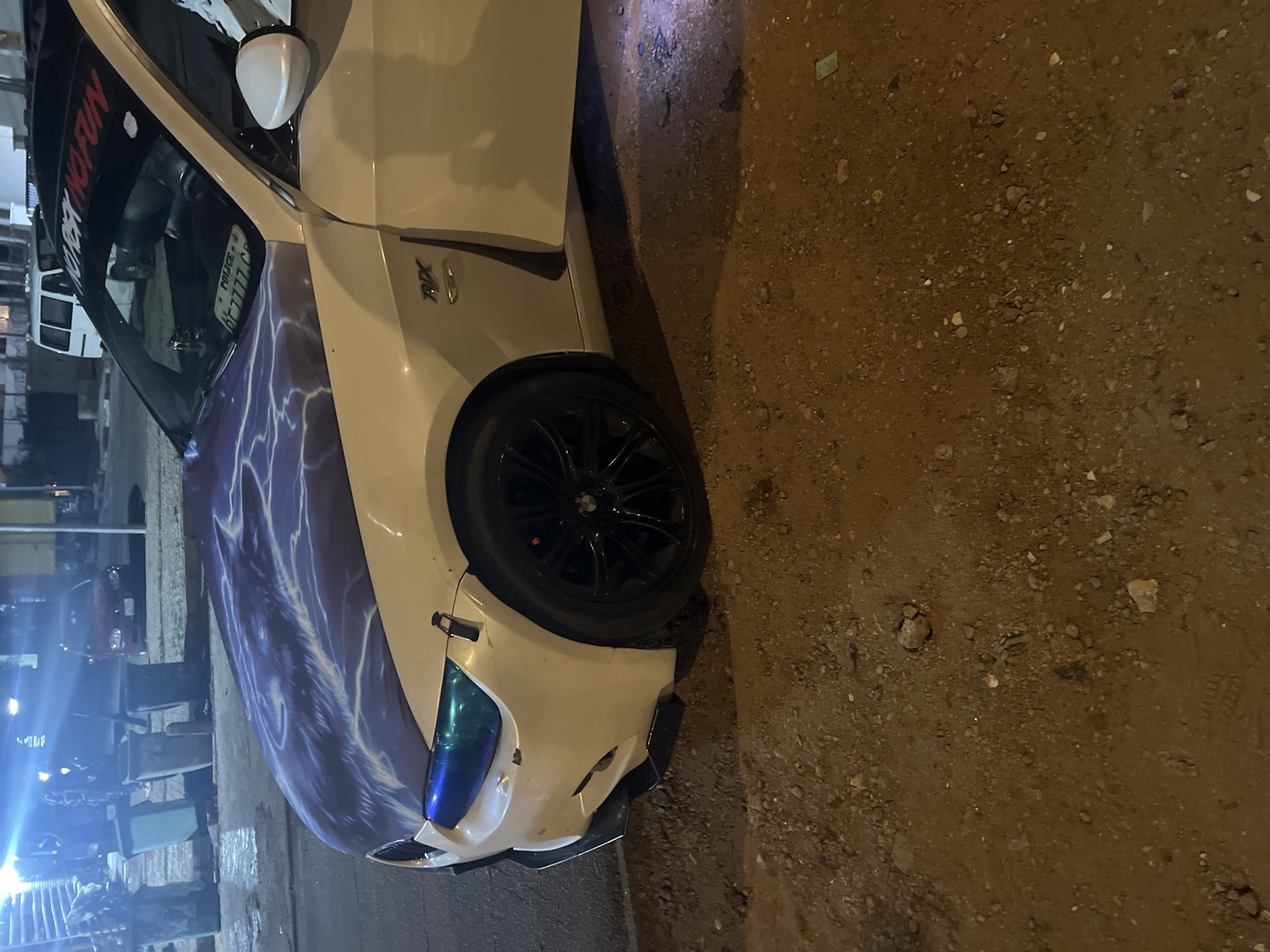 The image shows a modified white BMW RX7 parked on a street in Abuja, Nigeria. The car has a purple and white lightning bolt design on its hood and the windshield has a "NO RISK NO FUN" decal. It also has a blue-tinted headlight. The background shows a street scene at night with various buildings and streetlights. The ground is dirt and gravel.