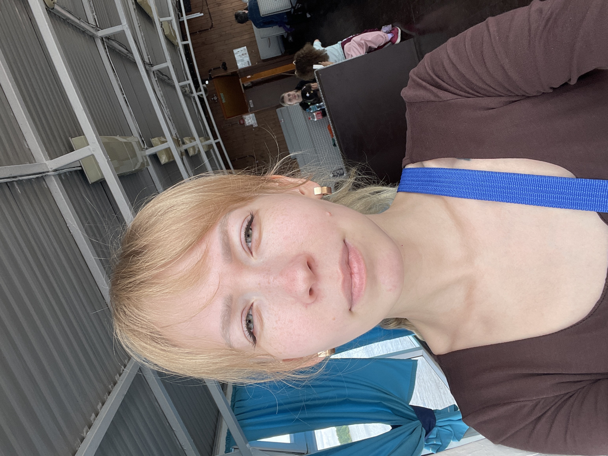 This selfie-style image, rotated ninety degrees clockwise, features a young woman with blonde hair, fair skin, and freckles, looking directly at the camera with a calm, neutral expression. Her eyes are light-colored, and she wears a small, gold-toned earring in her right ear. She is dressed in a brown long-sleeved top, with a distinct blue strap, possibly from a bag, visible across her chest.
The background reveals the interior of what appears to be a boat or ferry cabin, consistent with a leisure activity in Perm, Russia. The walls are made of light gray corrugated metal, supported by white structural beams, some of which have attached rectangular units, likely part of a ventilation or lighting system. Blue fabric, possibly curtains or draping, is visible near a window. Through the window, a bright body of water and distant green land are visible under daylight, suggesting good weather.
In the mid-ground, other individuals are present but blurred. One person with curly hair, dressed in a pink and white outfit, is seen sitting or resting on a dark surface, possibly a table or bench. The overall scene depicts a casual, daytime setting, likely during a scenic journey on the water. No visible text is identifiable in the image.