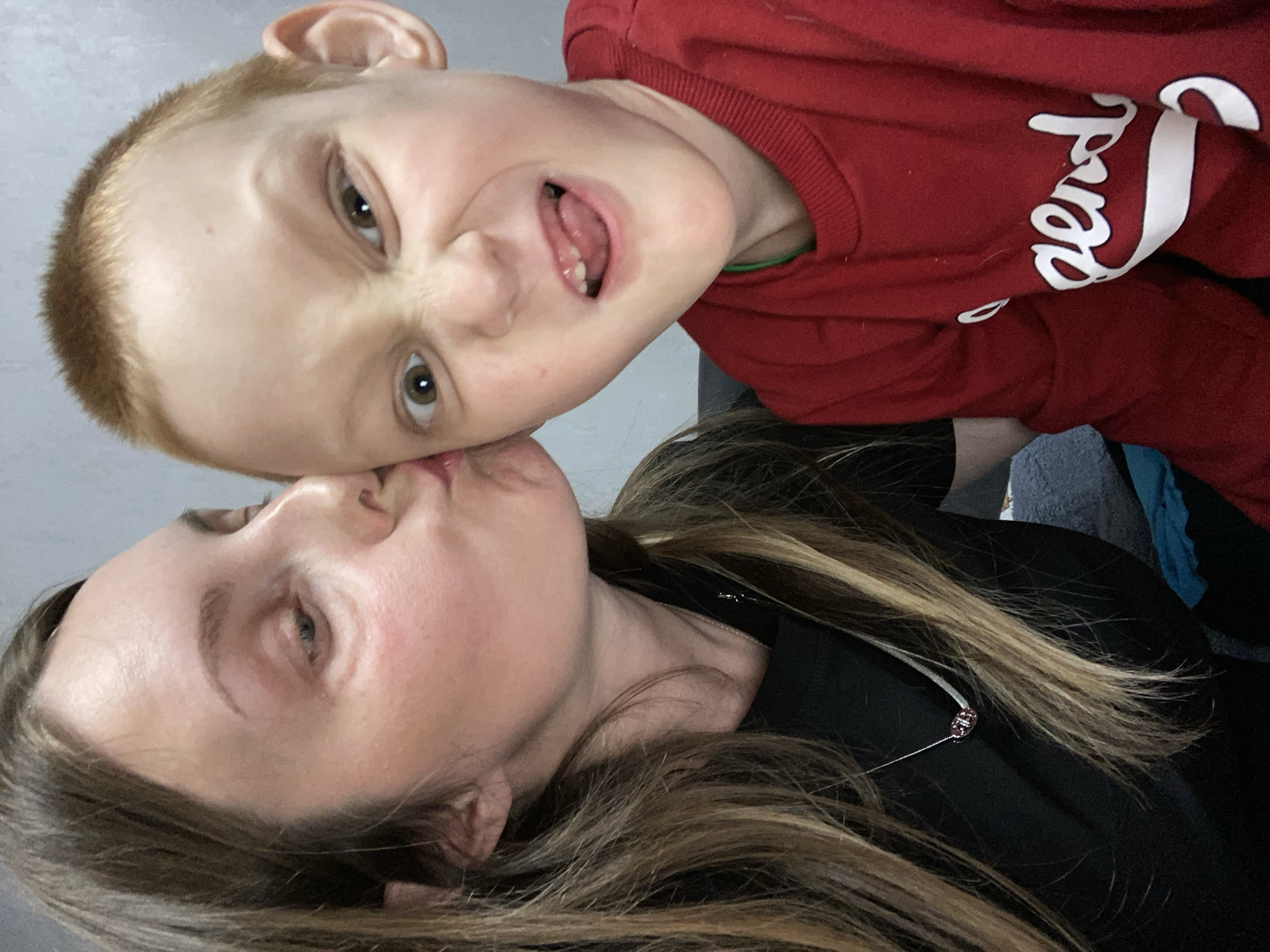 A woman and a young boy pose for a selfie. The woman, facing upwards, puckers her lips as if for a kiss. She has light skin and long brown hair. The boy, above her, makes a face with his mouth open and eyes wide. He has short red hair and wears a red sweatshirt with white lettering. The background is a blurry grey wall, suggesting an indoor setting. The photo is taken at an angle. Location details and city cannot be found in image.