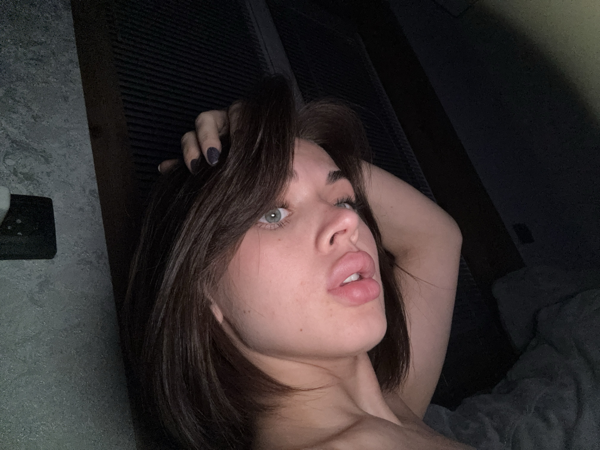 A young woman with short, dark hair and light-colored eyes is posing in a selfie. She is indoors. Her left hand is in her hair. The background consists of a textured wall and what appears to be a headboard of a bed. Лобаново, Russia is not discernible in the image.