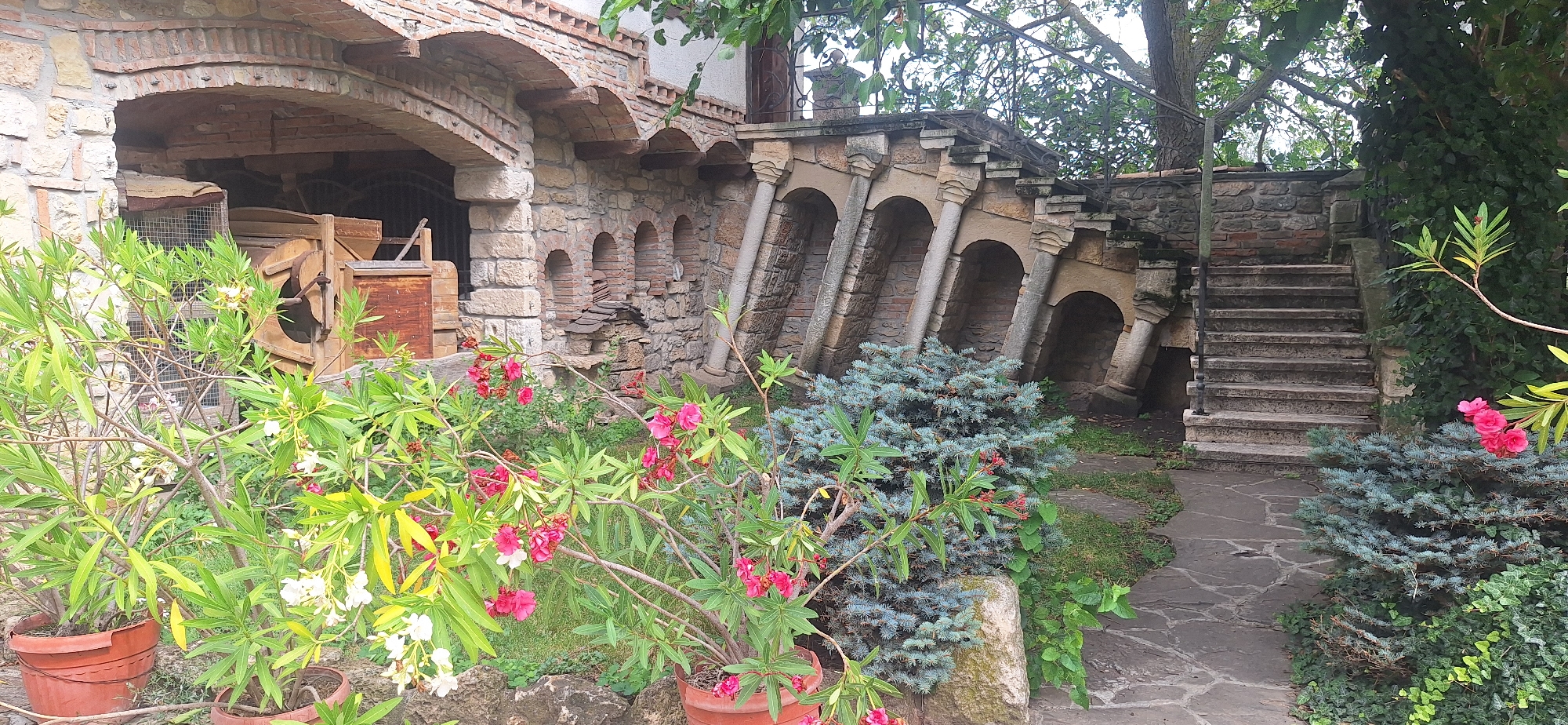The image depicts an outdoor scene at an old stone structure with arched openings and a staircase in Kalimantsi, Bulgaria. In the foreground, several potted plants with pink and white flowers are visible. To the left, a stone archway reveals machinery and wooden crates stored inside. The central part of the image features a stone wall with multiple arched niches supported by pillars. To the right, a stone staircase ascends alongside the wall, which is partially covered in ivy. The ground is paved with irregular stone slabs. The overall environment suggests a historical or rustic setting.