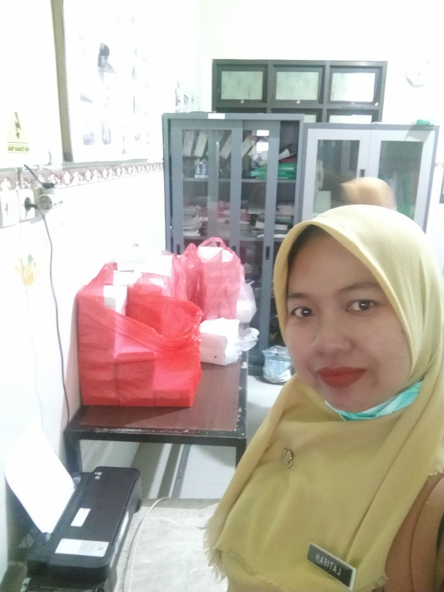 The image shows a woman wearing a yellow hijab and a light blue mask. She has red lipstick on and a name tag that reads "HABITAH" pinned to her hijab. In the background, there are several red plastic bags filled with white boxes, and two gray cabinets. To the left of the bags, a black printer with paper is visible. The overall scene appears to be an indoor setting, possibly an office or a store, given the presence of cabinets and office supplies. The lighting in the image is bright and somewhat washed out, indicating a possible overexposure or strong artificial lighting. There is no direct indication in the image that it is located in Trawas, Indonesia, but if this is assumed to be the case, then the scene depicts an everyday interior environment within that city.