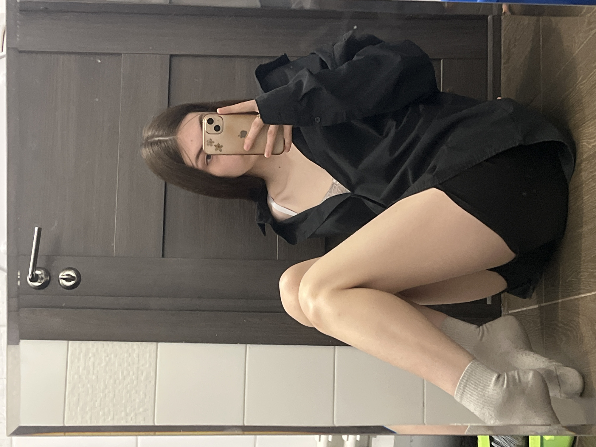 A woman is posing for a mirror selfie in what appears to be a bathroom. She is wearing a black oversized shirt, black shorts, and white socks. The woman is holding her phone in her right hand, partially obscuring her face. Her long brown hair is visible. The bathroom has gray cabinets with a brushed silver handle, and white rectangular tiles. The lighting is bright, seemingly from both natural and artificial sources. The photograph is taken in a tight space, likely reflecting the compact nature of urban bathrooms. It is impossible to locate the photograph within Saint Petersburg, Russia.