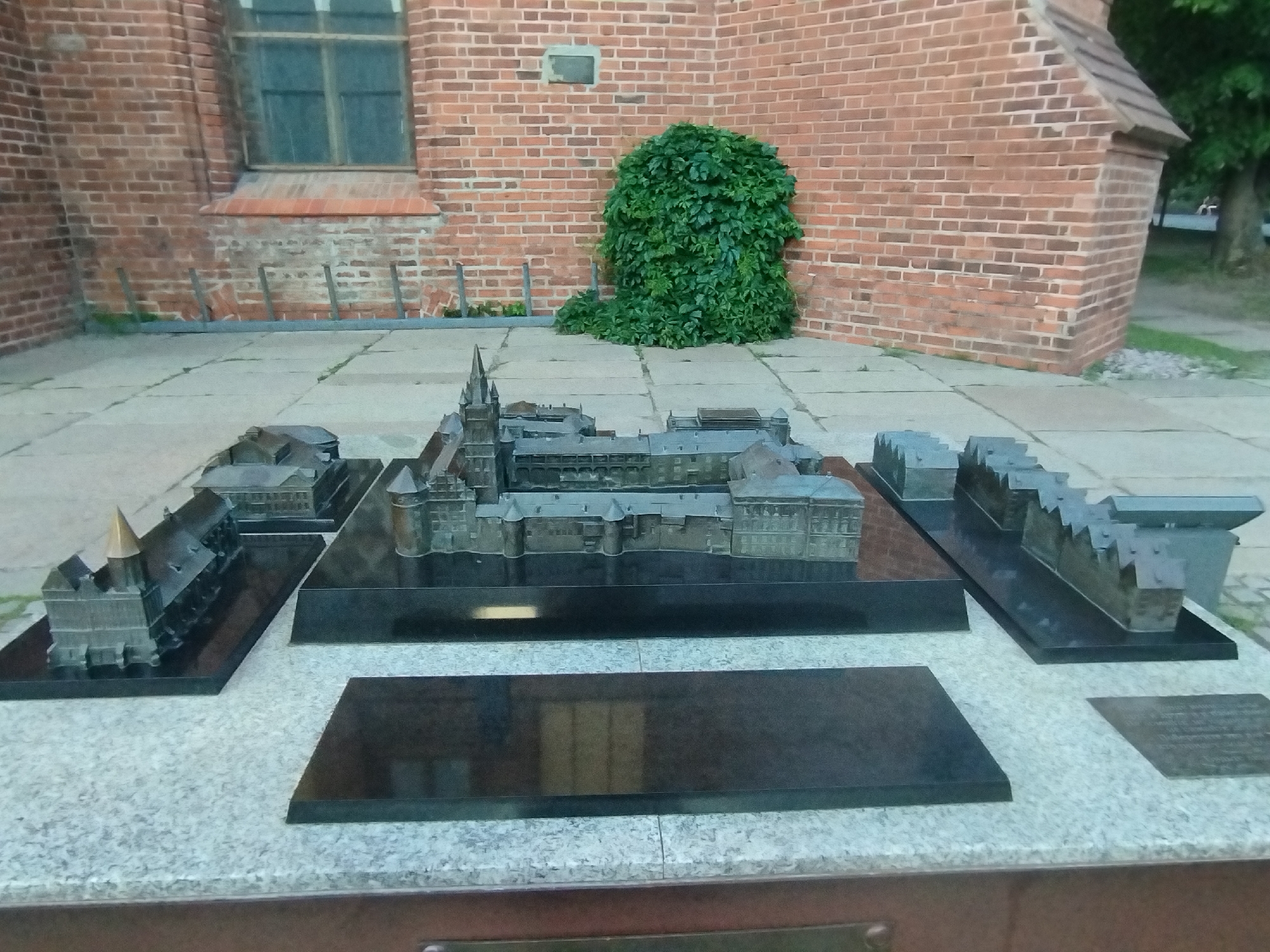 A detailed architectural model of a historical building complex is displayed outdoors on a granite pedestal. The model, constructed from dark metal, depicts multiple structures with varied rooflines, towers, and facades. It is arranged on raised, dark platforms that create a multi-leveled presentation. The background features a red brick wall of a building, a large green bush, and paved ground. The overall scene appears to be in a public space, possibly near the actual historical site the model represents, which is a representation of the Königsberg Castle, located in Kaliningrad, Russia.