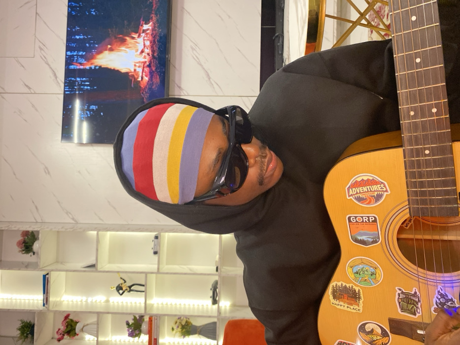 A person wearing a black hoodie, sunglasses, and a striped headband is positioned holding a guitar. The guitar is adorned with adventure and nature stickers. A television displaying a fire scene hangs on the wall. Behind the person is a lit white shelving unit with various decorations, including flowers and figurines. The wall has a white marble pattern.