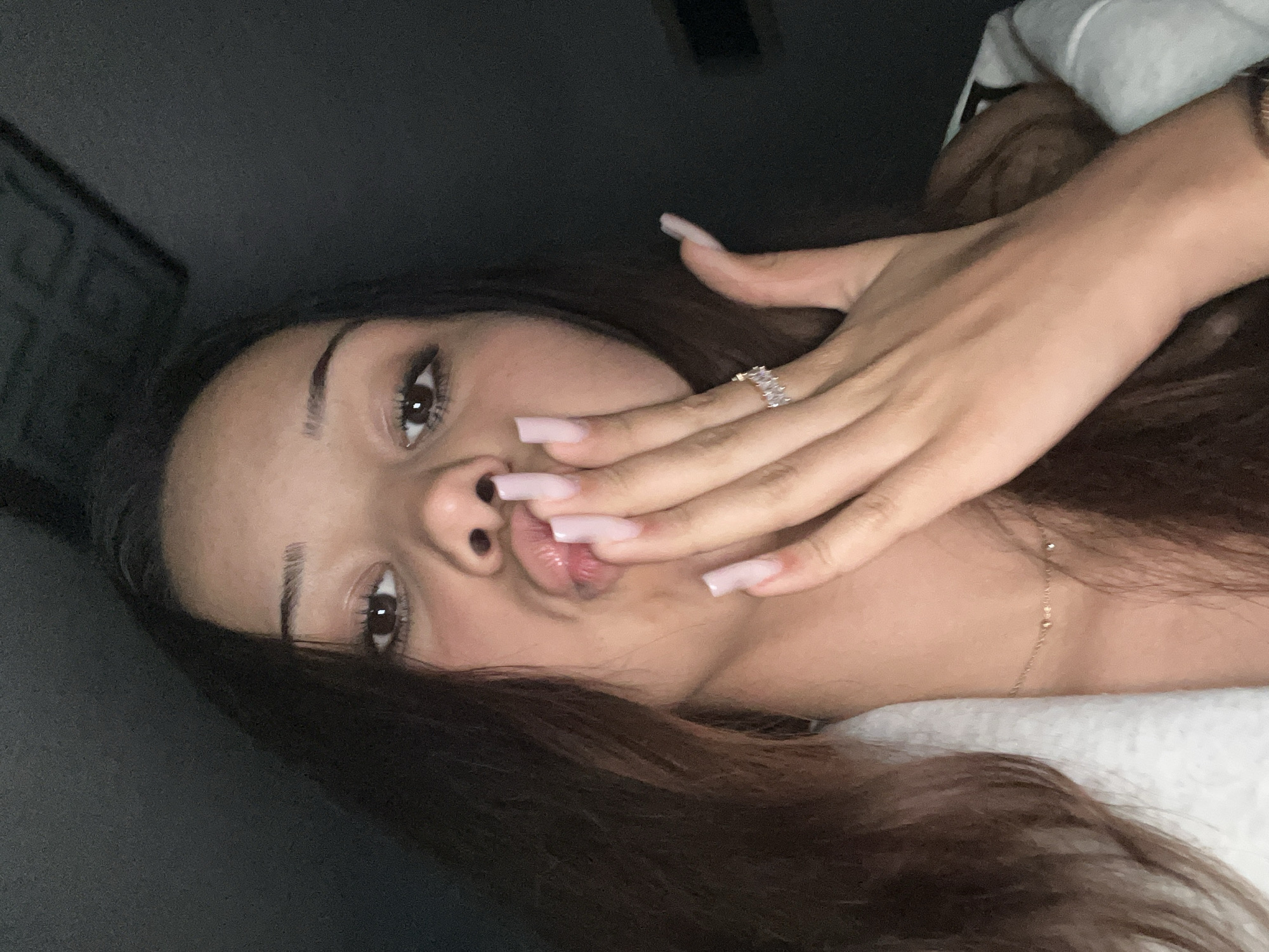 A young woman with long, dark brown hair, a medium complexion, and dark brown eyes looks directly at the camera. Her lips are pursed in a pouting gesture. Her right hand is raised, with pale pink, long, square-tipped fingernails partially covering the lower half of her face. A ring with multiple clear stones is worn on her right ring finger. A delicate gold-colored necklace with small beads is visible around her neck. She is wearing a light-colored garment, possibly white or light grey. The background is uniformly dark, indicating a low-light indoor environment. Location: Unknown, Unknown.