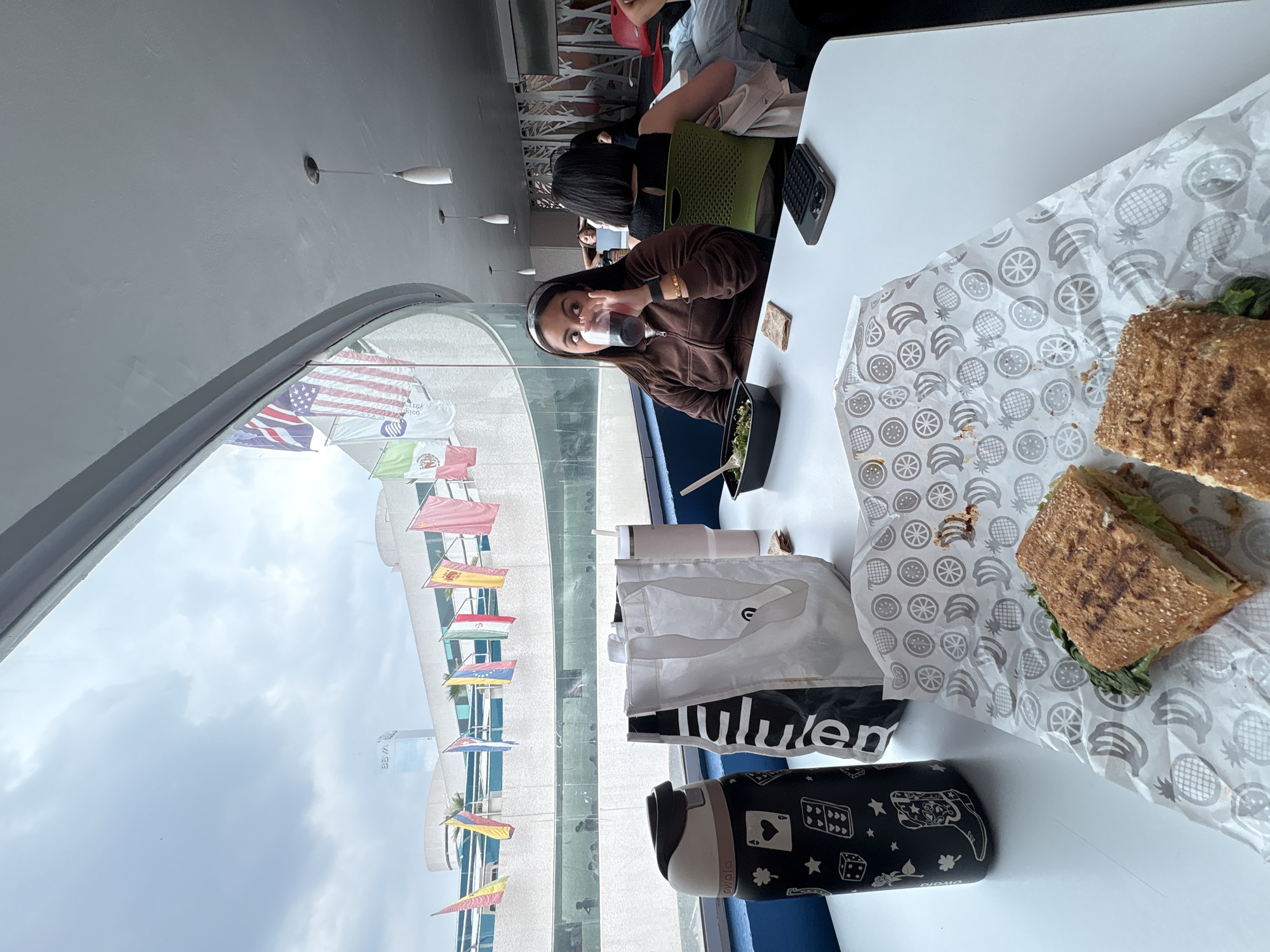 A woman sits at a table near a window in Puebla, Mexico. She wears a brown jacket and a face mask pulled down under her chin. On the table are a sandwich, salad, a Lululemon bag, and a water bottle. Other people sit at tables nearby. Flags are visible outside the window.