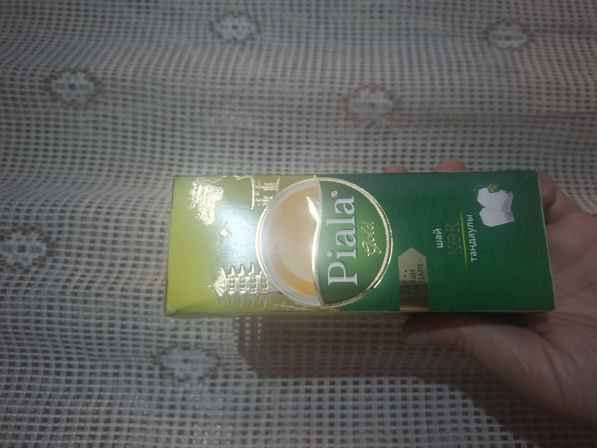 A close-up shot depicts a hand holding a green and yellow box of "Piala Gold" tea against a blurred patterned background. The box is rectangular with gold accents and features the brand name prominently. The tea is "KOK" and a picture of teabags is on the box. The location is likely indoors, and the specific city is not determinable from the visual information.