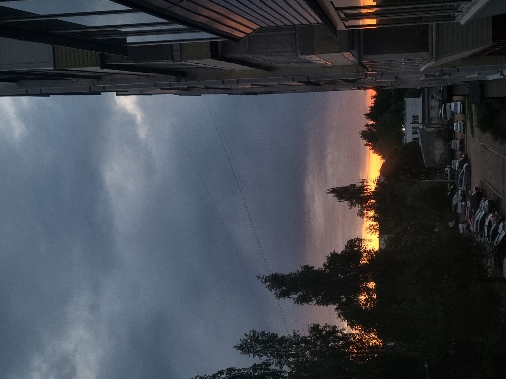 The image captures a sunset scene over an urban or suburban landscape in Chaikovsky, Russia, viewed from an elevated position. The scene is dominated by a wide, cloudy sky and the warm glow of the setting sun on the horizon.
In the upper part of the image, dark, angular structures of a building, likely the vantage point of the photographer, frame the scene. Some windows on the left side of this building reflect the vibrant colors of the sunset. Below this, the sky occupies the central and left portions of the image, appearing dark blue-gray with visible clouds, indicating an overcast evening. Faint, thin utility wires stretch horizontally across the sky.
Towards the right, the horizon is illuminated by the bright, intense colors of a sunset, featuring shades of vivid orange, yellow, and soft pink. A dense line of trees creates a dark silhouette against this bright backdrop, obscuring the direct view of the sun. Below the treeline, in the background, several buildings, including a prominent white structure, are visible, along with multiple parked cars on what appears to be a street or parking area. The overall atmosphere conveys the quiet transition from day to night in a residential area. No people are visible, and there is no discernible text in the image.
