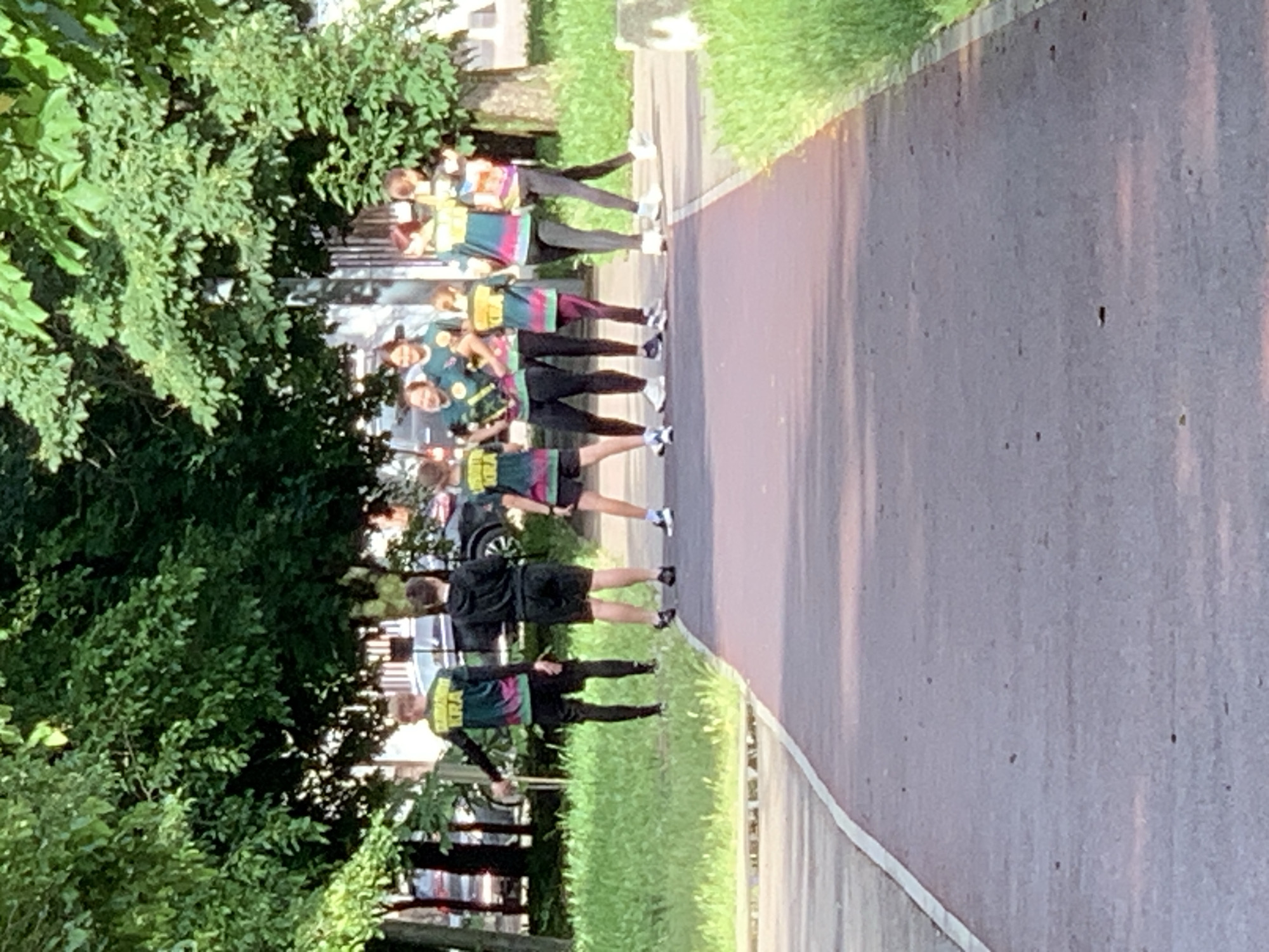 A group of people, seemingly part of a team, are standing on a paved path or road alongside a grassy area with trees and bushes. The individuals are wearing matching shirts with a colorful design and some are wearing black pants or shorts. The scene appears to be in an urban or suburban area, indicated by the presence of buildings and parked vehicles in the background. The perspective is from ground level, looking towards the group.