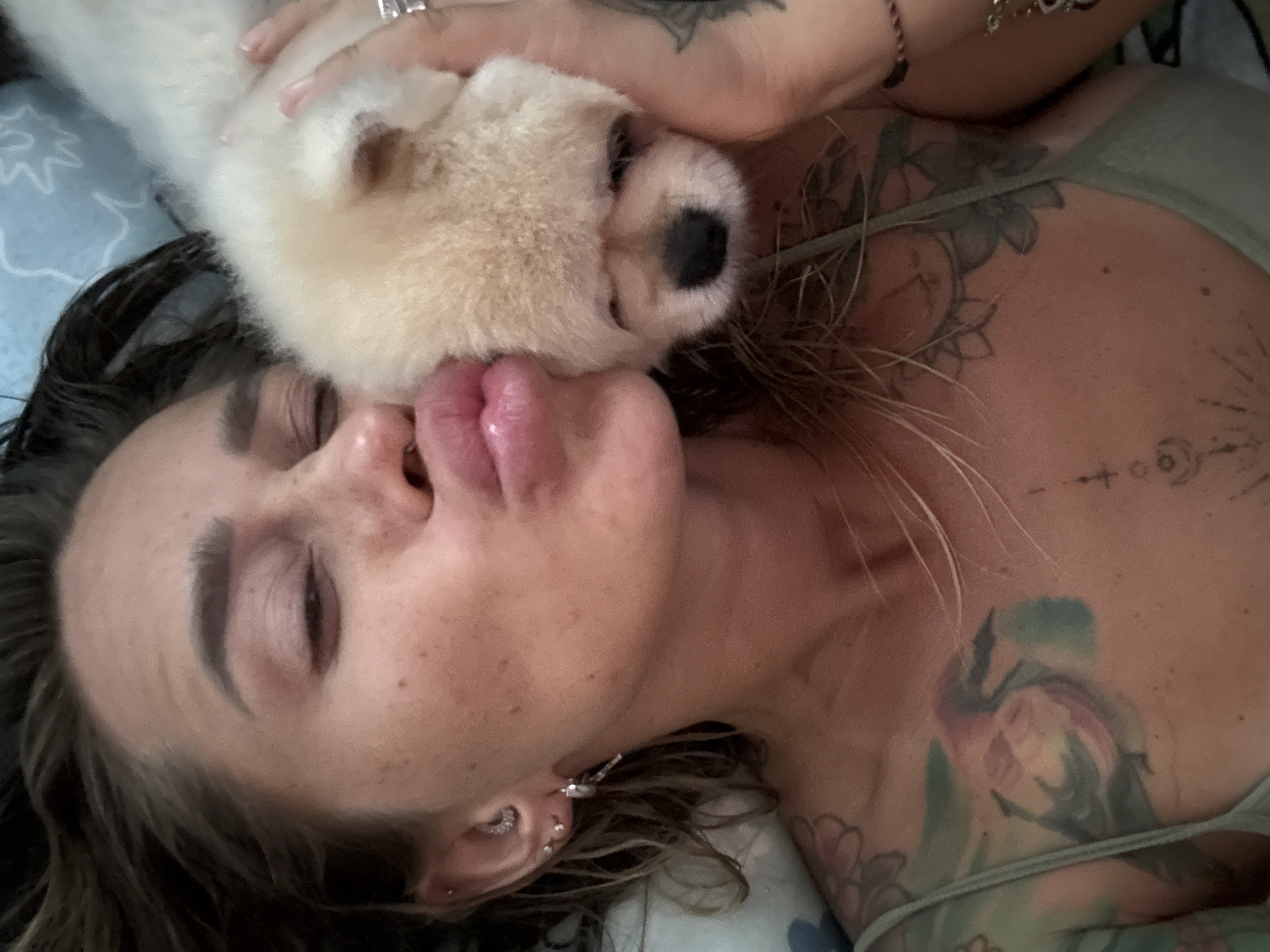 A close-up selfie shows a woman with her eyes closed, puckering her lips against the face of a small, fluffy, cream-colored dog. She is lying down, with her hair spread out. Her skin has freckles, and she is wearing several earrings and a ring on her left hand. Tattoos are visible on her chest and shoulder. The background appears to be a bed with a blue and white patterned sheet. The image was taken in Thep Krasatti, Thailand.