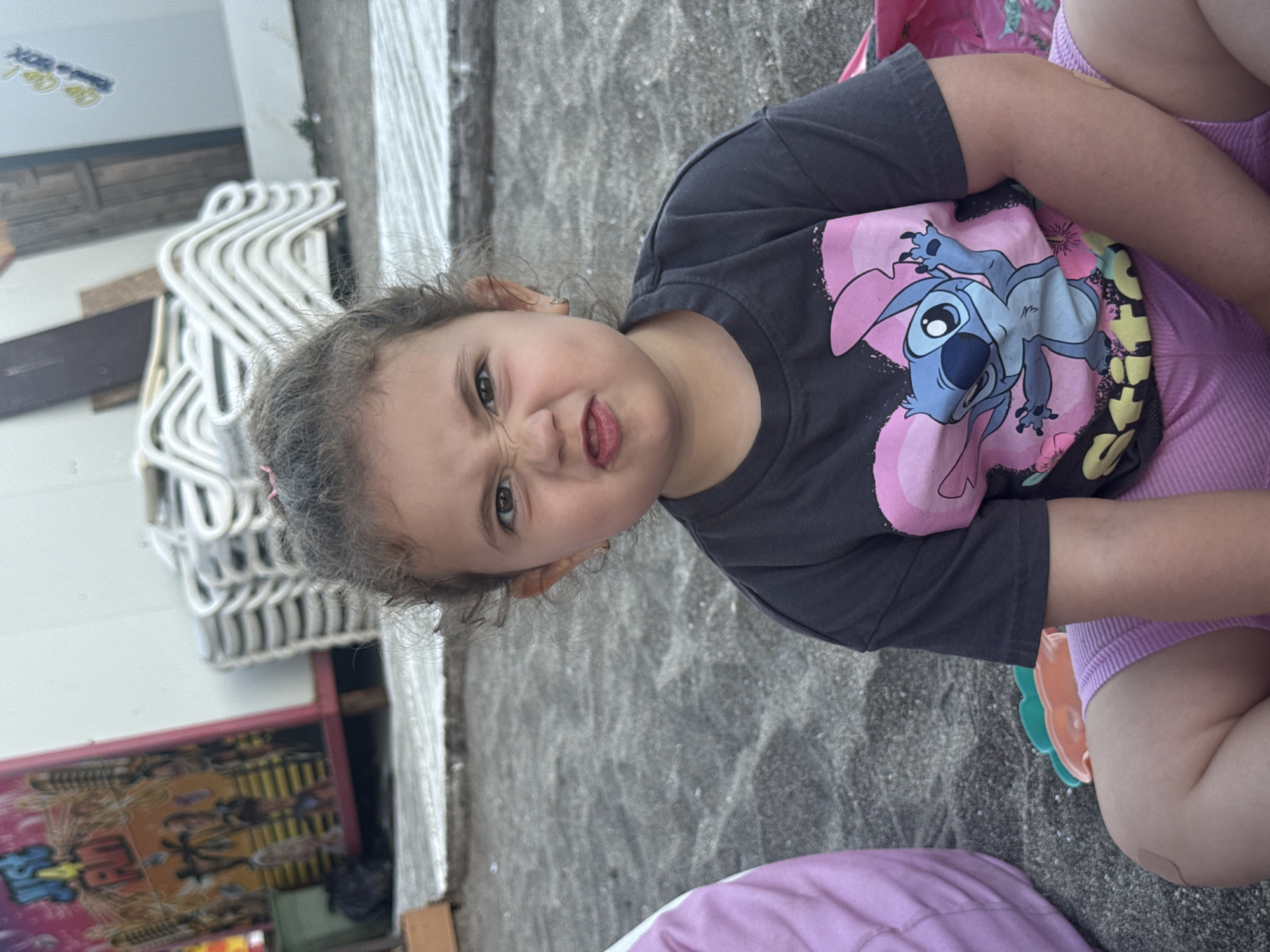 A young girl with curly brown hair is sitting on a dark sand beach. She is wearing a dark gray t-shirt with a pink and blue Stitch graphic and pink shorts. Her facial expression is one of playful defiance, with her nose wrinkled and mouth pursed. To her left is a large, plush purple cushion. In the background, there are stacks of white lounge chairs and a colorful carnival-style booth. The scene is set in Burgas, Bulgaria, likely at the beach or promenade area.