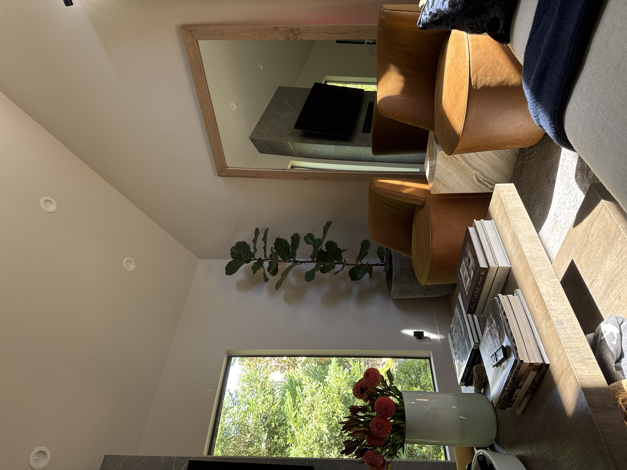 A sunlit, contemporary living room in Walnut Creek, USA, features two caramel-colored leather chairs positioned near a large, natural wood-framed mirror. The mirror reflects a modern fireplace with a dark gray marble finish and a mounted black television. A tall, potted fiddle-leaf fig plant stands to the left of the chairs. In the foreground, a light wood coffee table displays a stack of books, a remote control, and a white vase filled with red flowers. A large window to the left of the scene looks out onto lush green trees.