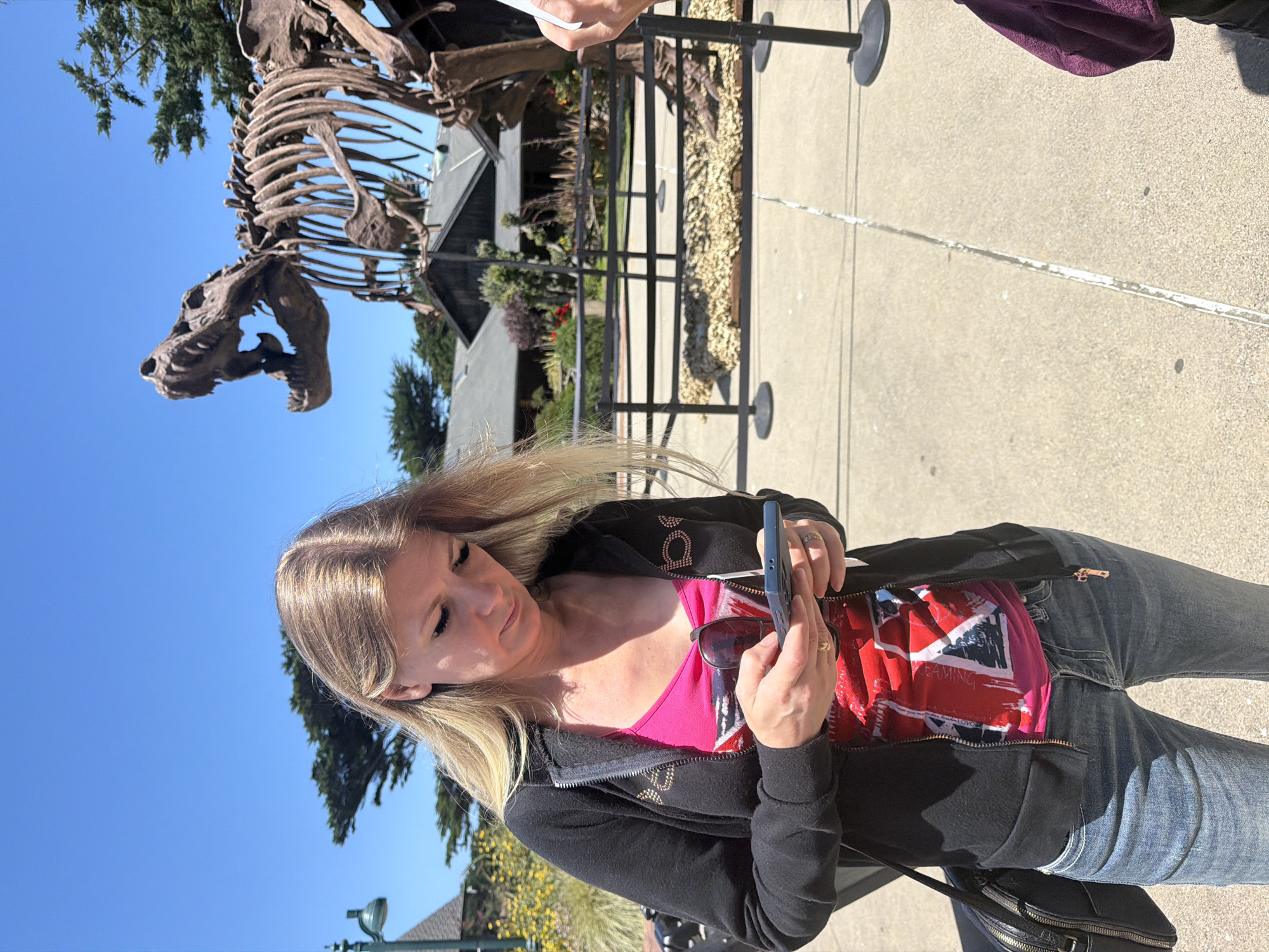 A blonde woman wearing a black zip-up hoodie over a pink tank top with a graphic print and blue jeans is looking down at her phone. She is holding a pair of sunglasses in her left hand. She has a black purse slung over her left shoulder. Behind her, a large dinosaur skeleton is on display outdoors. The background includes trees, buildings, and a blue sky. The scene appears to be a museum or outdoor exhibit. The location is likely the California Academy of Sciences in San Francisco, United States of America.
