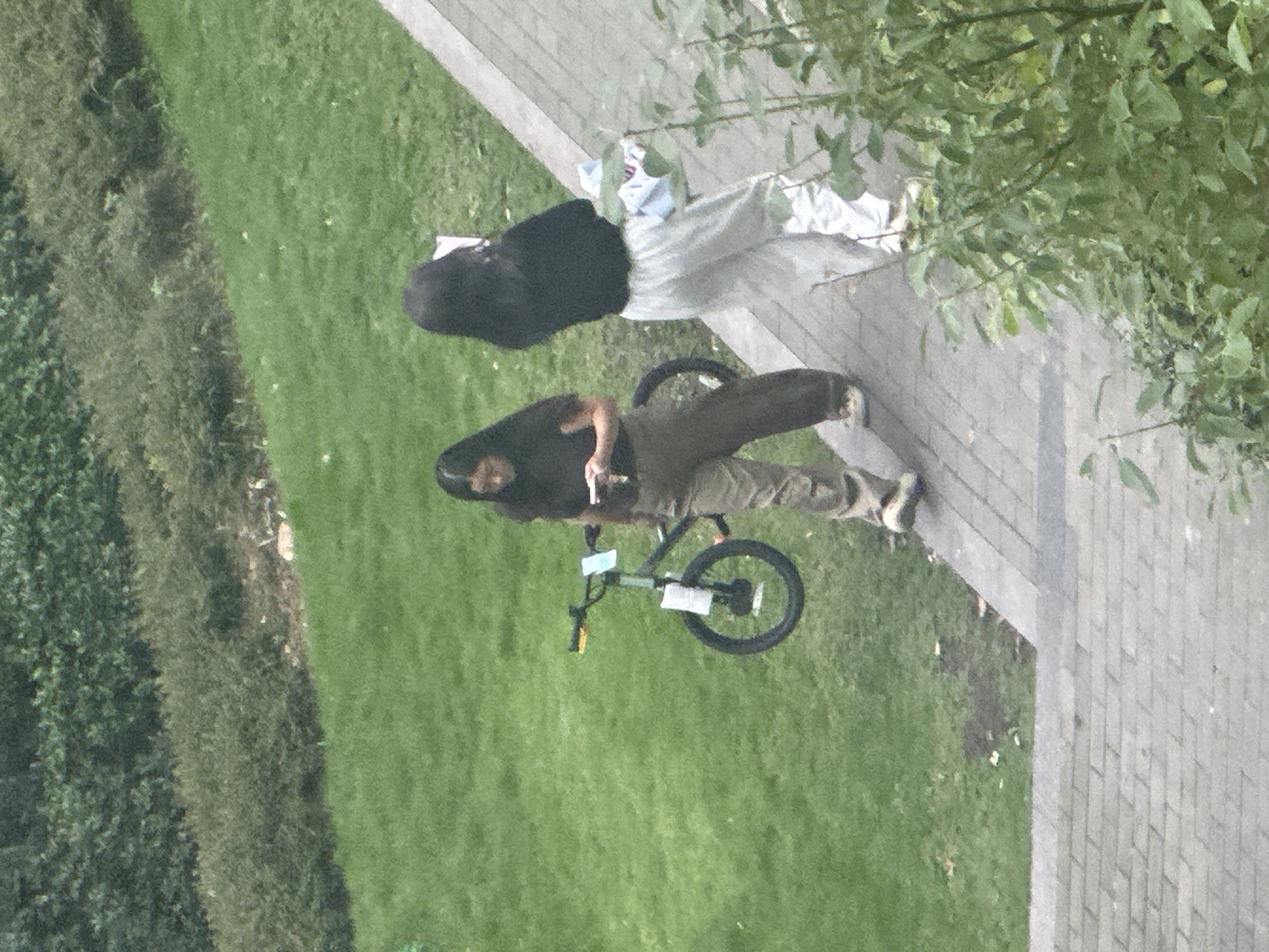 Two young women are walking on a paved path next to a grassy slope. One woman, on the left, is wearing a dark t-shirt and olive-colored pants and is holding a mobile phone. A bicycle, with two white papers attached to it, is positioned on the grass to her left. The second woman, on the right, is wearing a black long-sleeved shirt and light-colored pants and is carrying a bundle of folded clothes. The environment appears to be an outdoor setting with green vegetation and a paved walkway. The scene takes place in the Chongqing High-tech Zone, China.