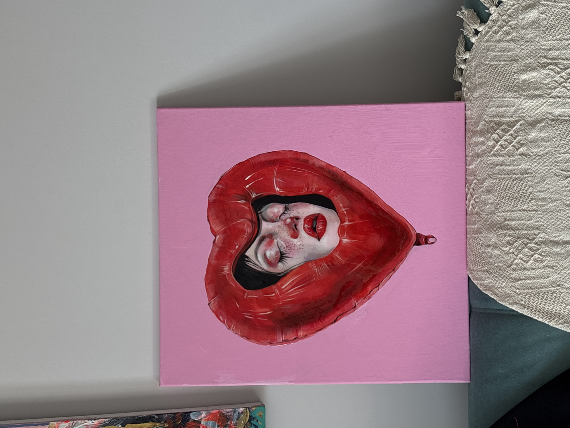An art piece, likely a painting on canvas, is displayed in an indoor setting in Krasnoyarsk, Russia. The canvas features a striking image of a stylized human face, appearing to be female, nestled within a large, glossy red heart shape. The face is depicted with closed eyes, long dark lashes, pronounced red lips, and scattered freckles across the nose and cheeks. Dark hair frames the top of the face, contrasting with the pale skin. The red heart, resembling an inflated foil balloon or a plush object due to its texture and sheen, prominently frames the face. The background of the painting is a solid, light pink, creating a vibrant pop-art or surreal aesthetic.
The painting is positioned upright, leaning against a light grey wall. To the right of the canvas, a beige or off-white textured throw blanket or pillow, adorned with tassels, rests on what appears to be a dark turquoise or blue-green upholstered sofa or armchair. A subtle shadow is cast on the grey wall above the painting, indicating the direction of light. A small portion of another colorful, abstract painting is visible at the very bottom edge of the frame, suggesting a collection of artworks. The scene evokes a calm, artistic atmosphere. No visible text is present in the image.