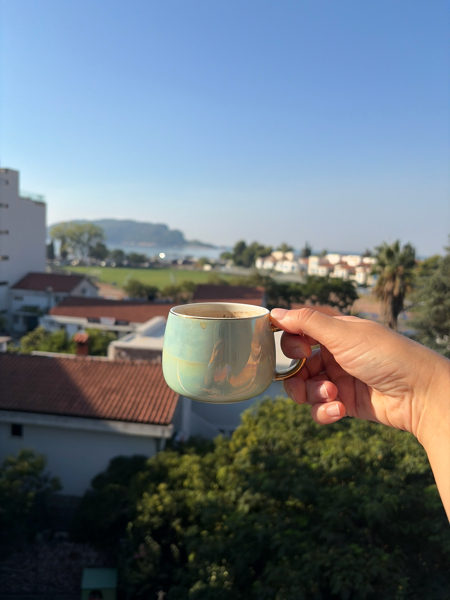 A hand holds a small, iridescent teal mug filled with coffee. The mug has a golden rim and handle. In the background, a blurred landscape of Budva, Montenegro, unfolds under a clear blue sky. Buildings with terracotta roofs are visible, interspersed with lush green trees and palm trees. In the distance, a body of water and a hilly landmass are faintly discernible. The scene is illuminated by bright sunlight, casting soft shadows.