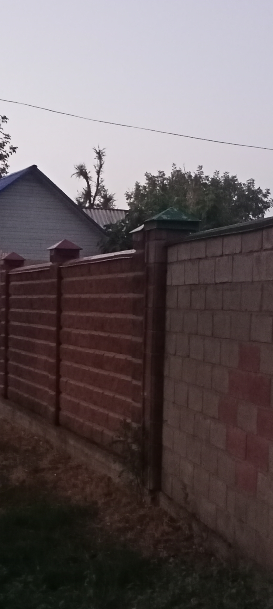 A residential property fence is visible in Sokuluk, Kyrgyzstan, under an overcast sky. The fence is constructed from horizontal red brick-patterned panels on the left and lighter colored brickwork on the right, meeting at a vertical concrete pillar topped with a green cap. A second fence section with red conical tops is visible behind the primary fence on the left. A portion of a building with a blue roof and siding is partially visible behind the fence on the far left. Trees and foliage are present behind the fence and building. The ground in front of the fence consists of dry grass and some scattered debris. A wire is strung across the top of the frame.