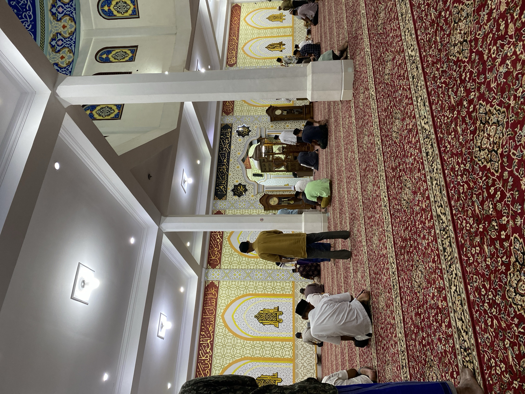 The image depicts the interior of a mosque, likely in Lhokseumawe, Indonesia, during a prayer service. Several men are seen sitting on a red carpet with intricate patterns. The walls are adorned with Arabic calligraphy and decorative yellow and white geometric designs, with arched recesses featuring ornate golden motifs. The ceiling is white with modern recessed lighting fixtures. In the background, a tall wooden clock and an ornate wooden structure, possibly a minbar or a decorative element, are visible. The atmosphere appears serene and reverent as the individuals engage in prayer.