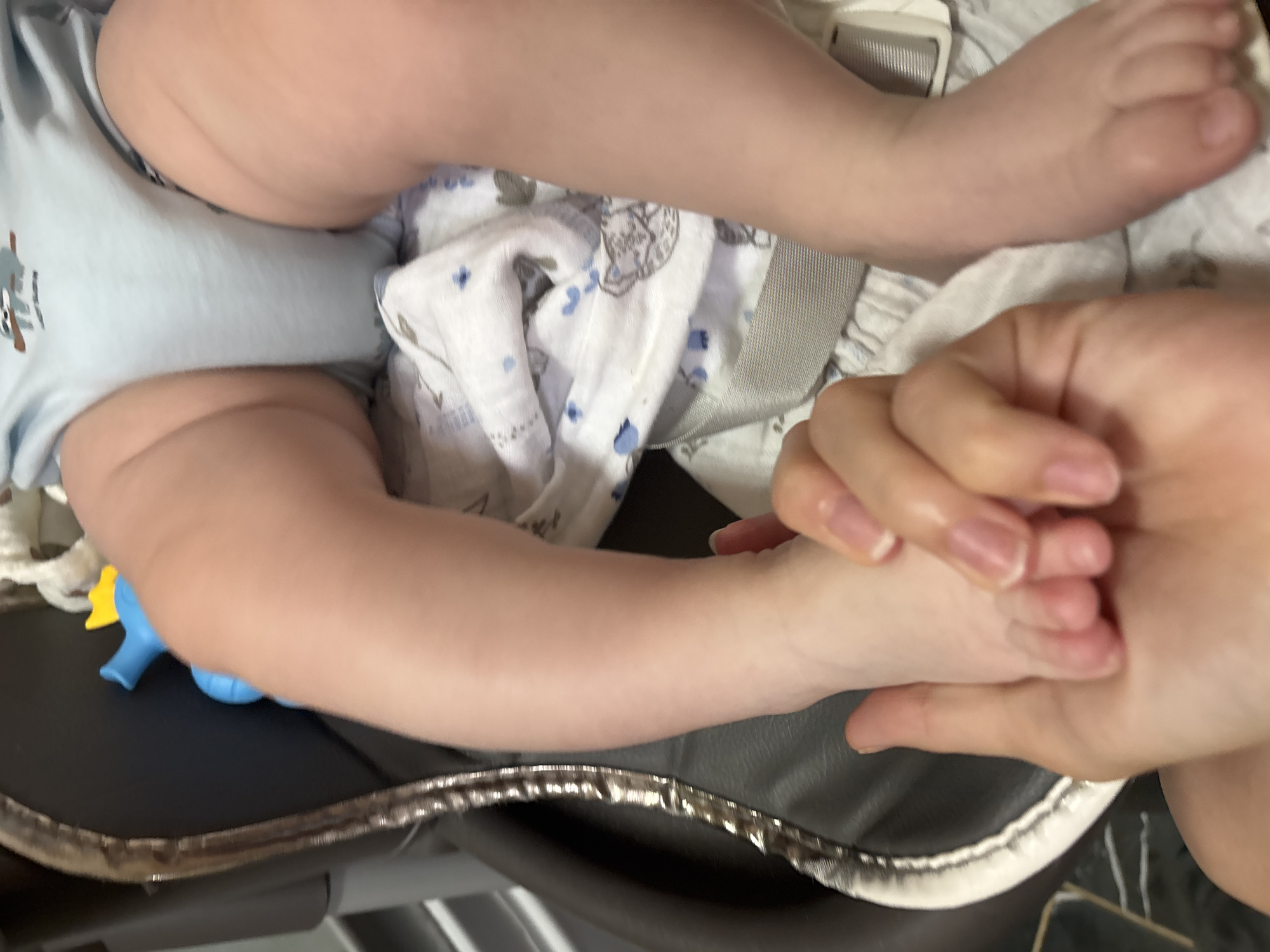 The image, taken in Razdolye, Russia, presents a close-up, high-angle view of a baby's lower body, partially obscured by an adult's hand. The baby is wearing a light blue bodysuit featuring a faded, cartoon-like grey and white animal design, possibly a bear holding an object resembling a shovel or stick. A white muslin blanket with scattered blue and grey patterns is draped around the baby's torso. An adult's hand gently cradles the baby's left foot, suggesting an act of care or affection.
The baby's bare legs and feet are visible, resting on a dark, possibly padded surface, which appears to be part of baby equipment like a stroller, car seat, or playpen, characterized by a metallic or reflective trim along its edge. A small, blue and yellow toy is partially visible near the baby's right arm. The scene is brightly lit, likely indoors, with no discernible time of day. No other specific location cues or visible text beyond the animal on the bodysuit are present. The interaction between the adult hand and the baby's foot conveys a sense of tenderness and connection.