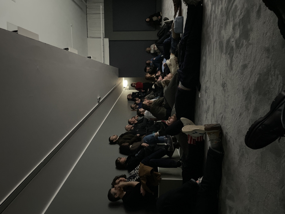 The image captures an indoor scene, likely within an art gallery, museum, or an immersive exhibition space. The primary visual element is a group of approximately 25-30 people, mostly adults, positioned in various states of repose and attention across a dark grey carpeted floor and low, light grey tiered steps. The image is originally rotated 90 degrees clockwise; when viewed upright, the people are facing towards the front of the room, looking upwards.
Many individuals are seen with their heads tilted back, gazing intently towards an unseen projection or display located above them, possibly on the ceiling or a very high wall. Some are lying fully on their backs on the floor or steps, while others are seated with their heads craned upwards. Their postures suggest an immersive viewing experience.
The room's aesthetic is minimalist and modern. The left wall is a plain, light grey, with a continuous strip of bright white light running horizontally along its base, illuminating the lower section. The floor where the majority of people are gathered is dark grey carpet. In the background, there's a darker wall section and what appears to be a slightly elevated area where a few more attendees are situated, including one person wearing a red top. Further back and higher up, a lone individual is visible, also looking upwards.
The overall lighting is subdued, with artificial light sources creating a focused ambiance, typical for such viewing experiences. Attendees are dressed in casual to smart-casual attire, including jackets, sweaters, and jeans. No discernible text is visible in the image. The atmosphere appears quiet and contemplative, with everyone focused on the unseen central display.