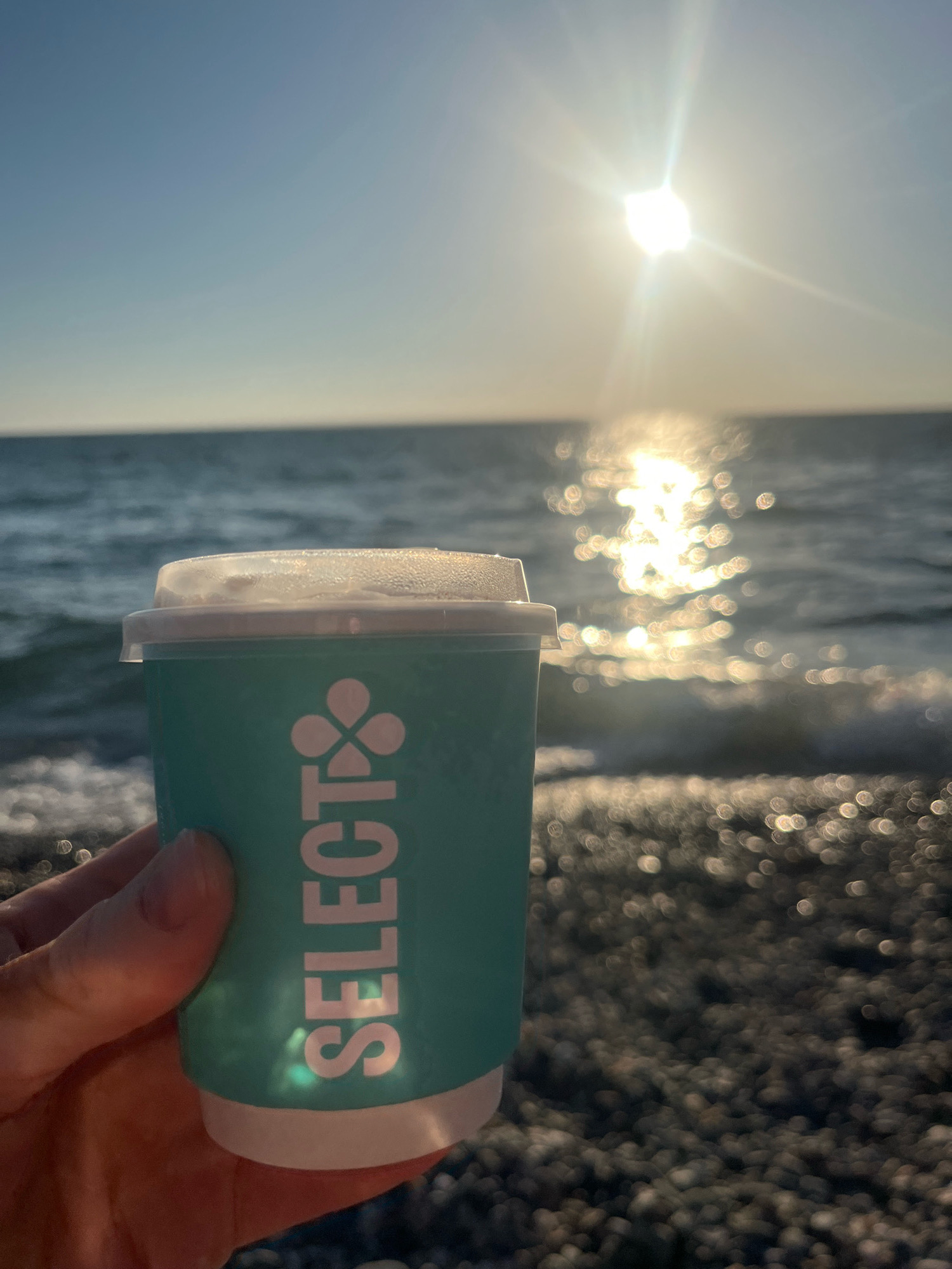 A hand holds a teal paper cup with a white lid. The cup has the word "SELECT" printed vertically in white capital letters, along with a white clover leaf logo. The cup is in the foreground, with a blurred background of the sea and a pebbled beach under a bright, sunny sky. The sun is high in the sky, creating a strong glare on the water. The image appears to be taken at sunset, given the warm light and the angle of the sun. The sea is calm, with small waves lapping at the shore. The pebbles on the beach are dark and appear wet. The overall impression is one of a peaceful and serene moment. This scene could be at any beach location with similar characteristics.