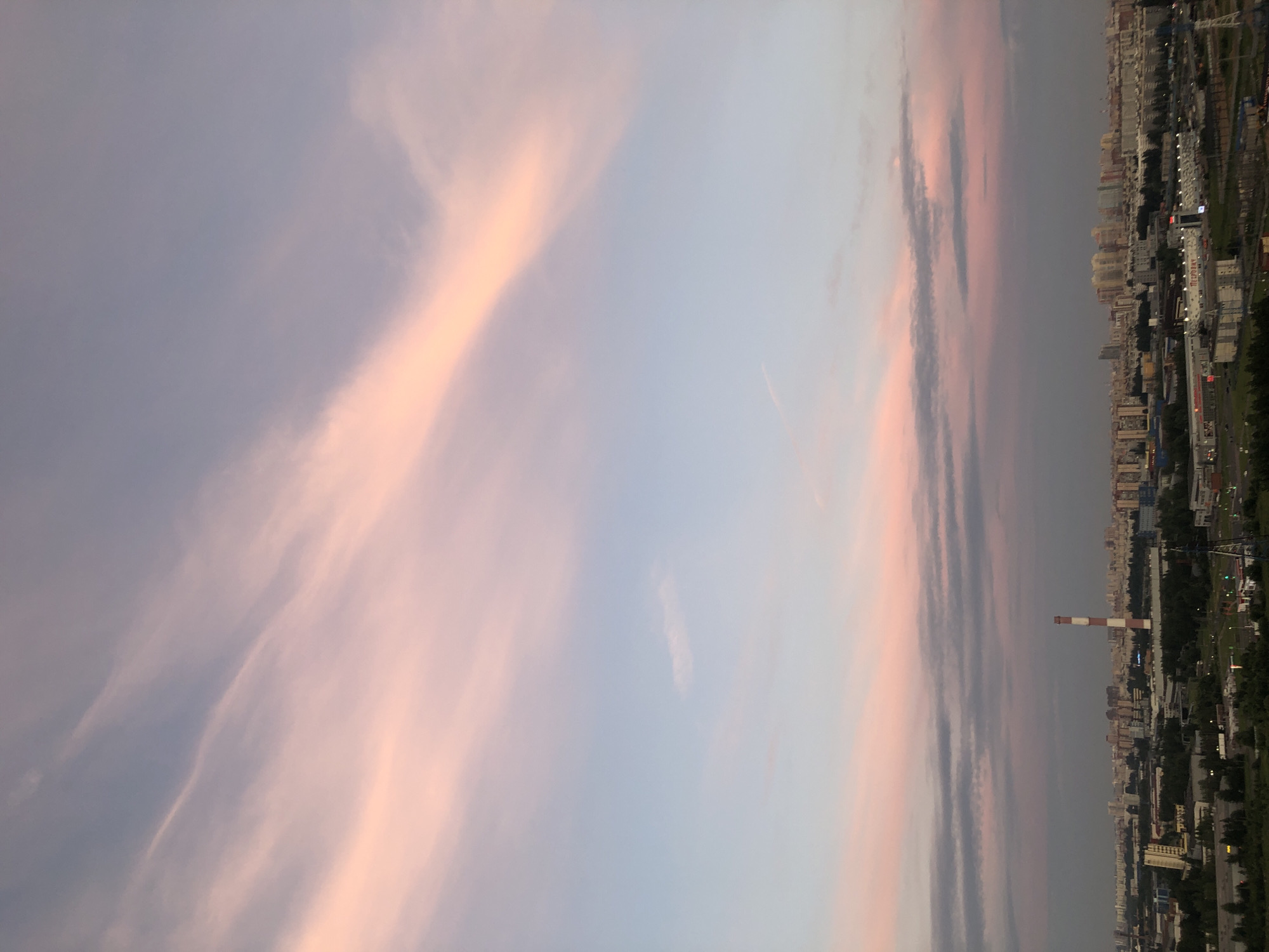 An aerial, full shot captures a cityscape at dusk under a pale pink and blue sky. The sky is filled with wispy clouds tinged with soft hues of pink and orange, characteristic of a sunset. Below the sky, a sprawling urban landscape stretches across the horizon. The city is characterized by a mix of residential buildings, industrial structures, and patches of green trees. A prominent red and white striped smokestack stands tall amidst the urban sprawl, suggesting an industrial or power generation facility. The overall atmosphere is tranquil, illuminated by the fading light of day. The exact location is unknown, but it depicts a dense urban environment with a visible industrial presence.
