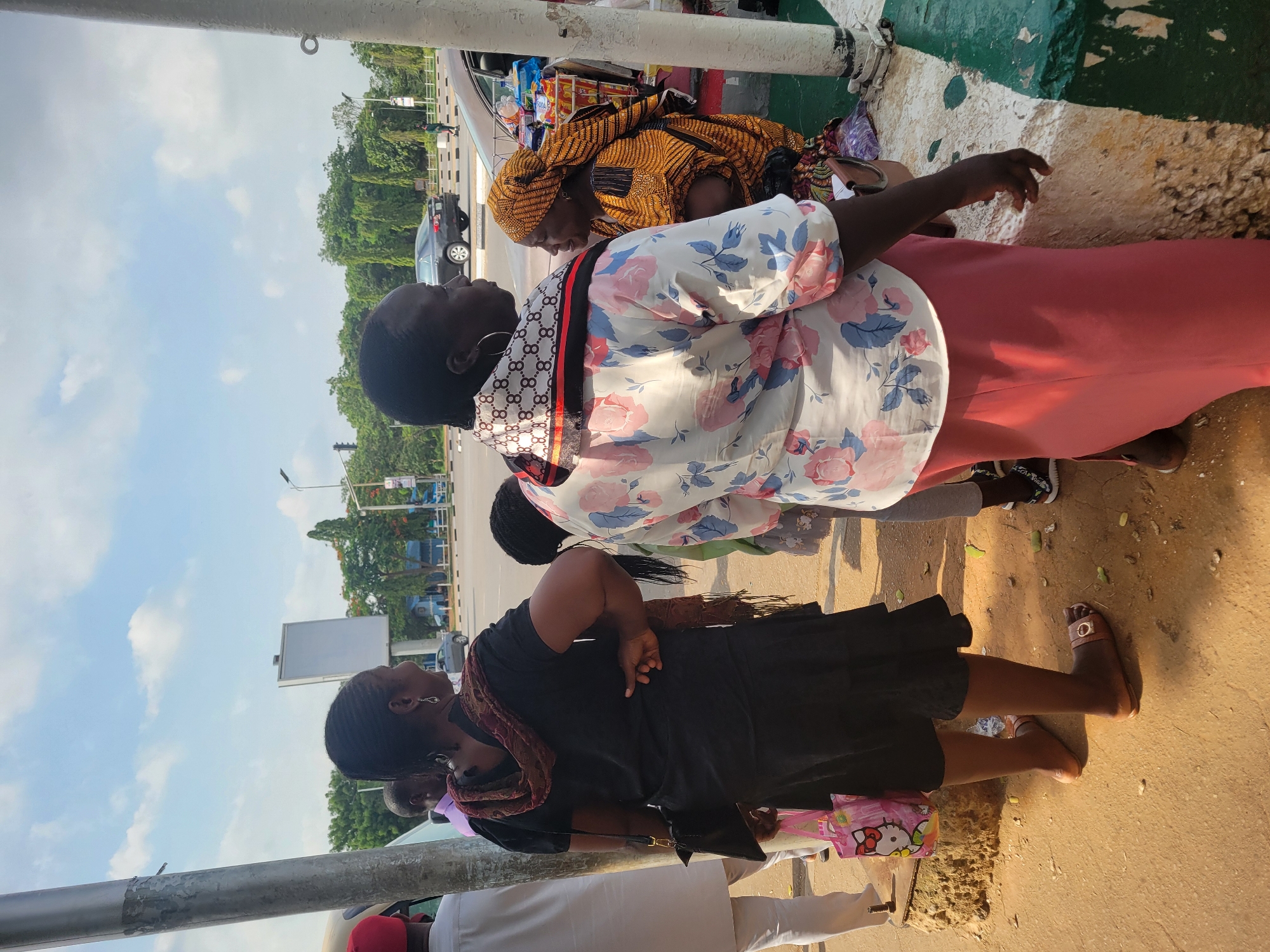 The image depicts a bustling street scene in Abuja, Nigeria, bathed in bright daylight under a partly cloudy sky. The photograph is oriented sideways, showing individuals gathered on a roadside or sidewalk adjacent to a multi-lane road lined with green trees.
In the mid-ground, a woman wearing a white short-sleeved top adorned with pink and blue floral patterns and a long peach-colored skirt stands. She has a distinctive black and white patterned neck scarf or bandana. To her left and slightly behind, another woman is dressed in a black top and skirt, accessorized with a dark, patterned scarf and carrying a pink bag with "Hello Kitty" written on it. Further to the right, a woman dressed in a vibrant yellow and black traditional patterned outfit with a matching headwrap appears to be a vendor. She is positioned next to what looks like a makeshift stall or the open trunk of a vehicle, displaying various packaged goods such as snacks and drinks. Partial figures of other individuals are visible in the foreground and background.
The background reveals a multi-lane road with a few cars, suggesting traffic. A street light and a white billboard are also present, indicating an urban setting. A large grey metallic pole is visible on the left side of the frame. The ground beneath the people consists of a mix of paved and dirt surfaces. The scene portrays everyday life, likely a market area or a busy transit point, with people engaging in commerce or waiting. Shadows indicate bright sunlight, suggesting a mid-morning or afternoon time.