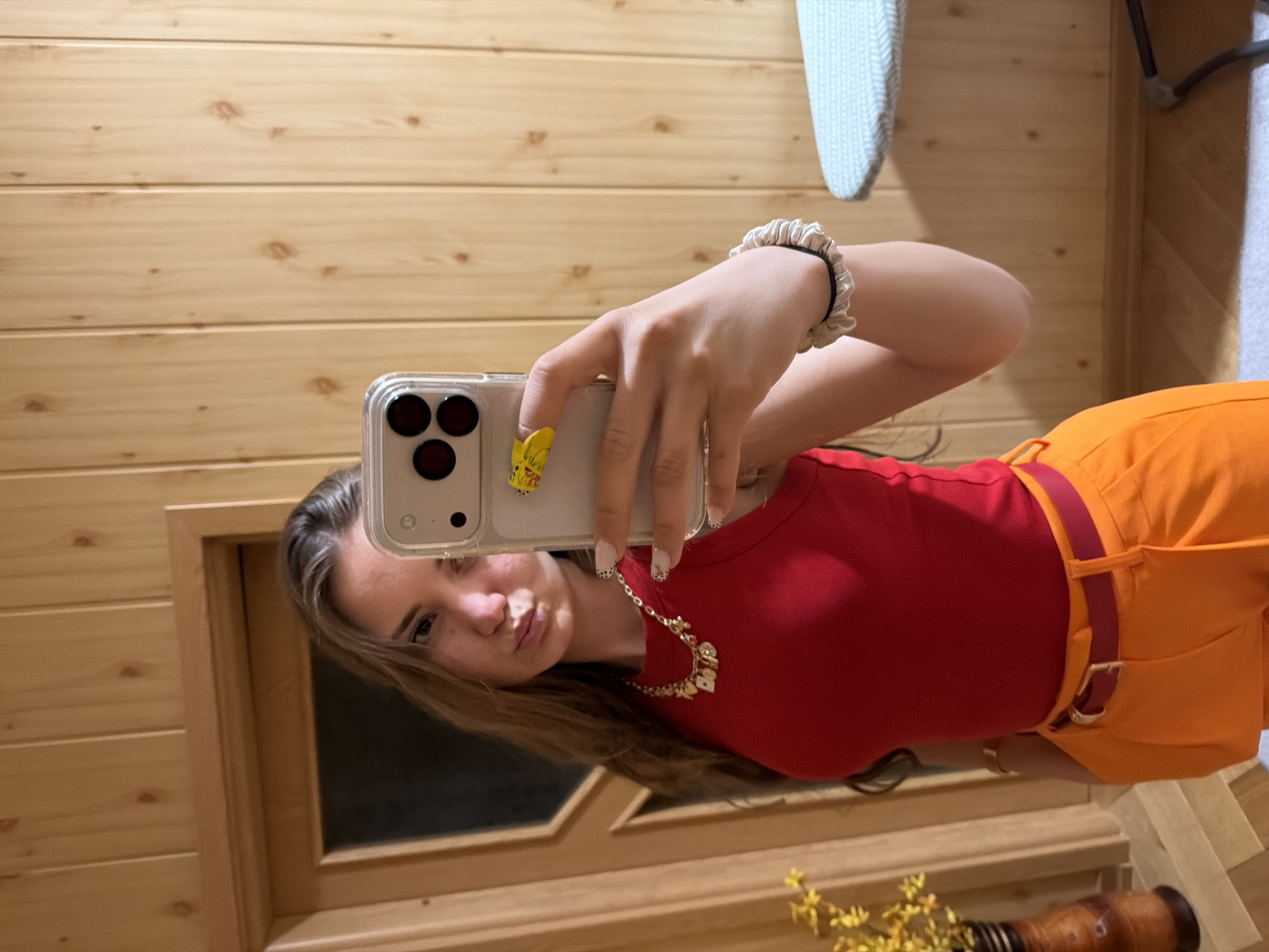 A young woman is taking a selfie in front of a mirror. She is wearing a red top and orange pants with a red belt. She is holding a phone in her hand, which has a yellow cartoon character attached to it. She also has a bracelet on her wrist and a necklace around her neck. The background is a wooden wall, likely paneling. It is impossible to determine from the image whether the location is Buhovo, Bulgaria.