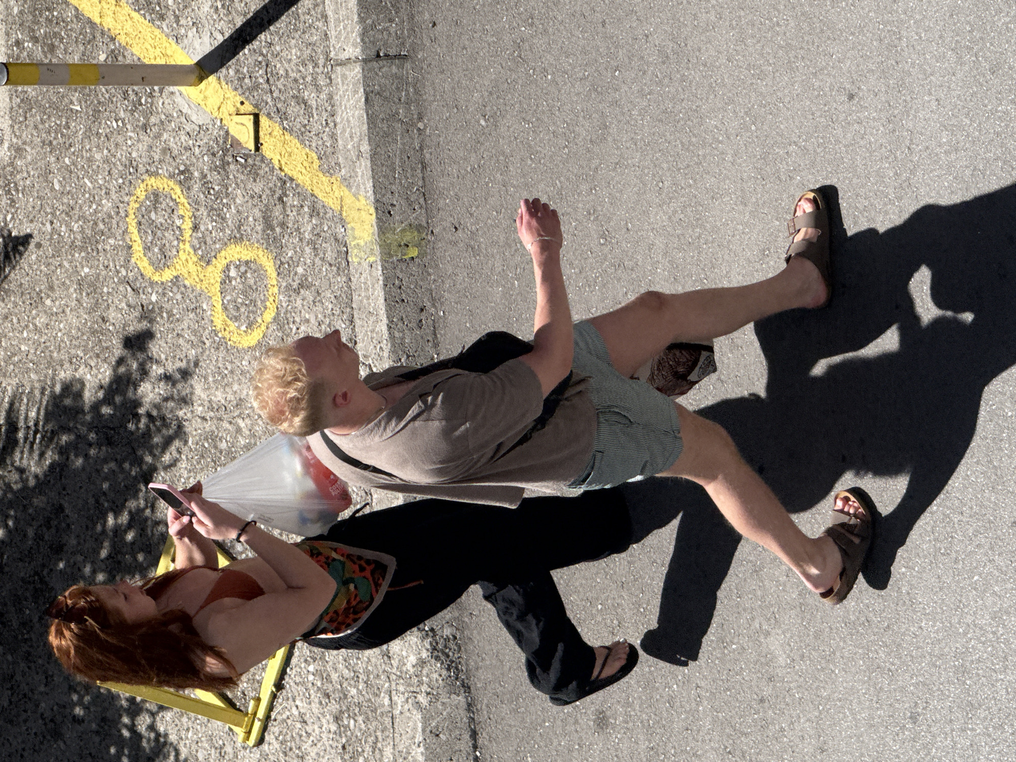 This overhead image, taken in Budva, Montenegro, shows two individuals walking on a grey asphalt surface under direct sunlight, casting prominent shadows. A young woman with long, reddish-brown hair and sunglasses on her head, wearing an orange bralette, black wide-leg pants, and black flip-flops, is holding a pink smartphone with both hands and carrying a transparent plastic bag containing items with red packaging. A patterned scarf is tied around her waist. To her right, a young man with short, light blonde hair is walking, wearing a taupe t-shirt, light blue and white striped shorts, and brown two-strap sandals, with a dark shoulder bag strap visible. The asphalt features a bright yellow diagonal line running alongside a concrete curb, and a yellow figure-eight symbol painted on the pavement. A yellow triangular metal barrier is positioned in the upper left, and a yellow and white striped bollard stands near the curb in the upper right.