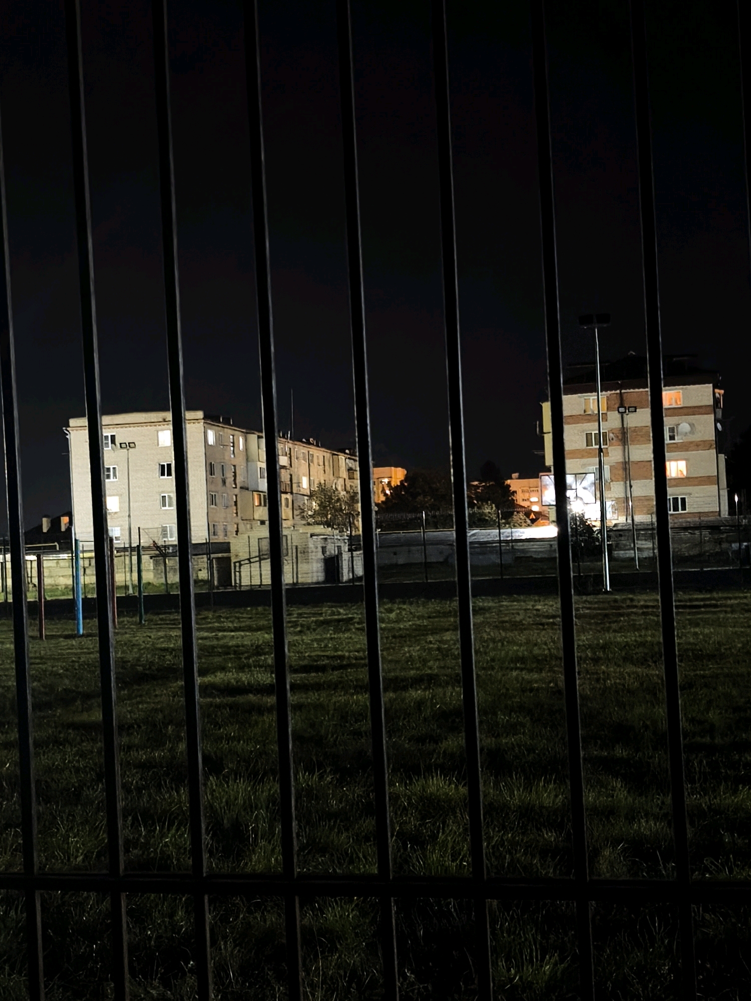 The image, taken at night, shows a view through a metal fence onto a grassy field with buildings in the background. The field appears to be a park or sports area, with some colorful poles visible on the left. Behind the field, there are apartment buildings illuminated by internal lights and exterior lighting. One building on the right has a distinct orange and white striped facade. The sky is dark, indicating nighttime. The scene is set in Karachayevsk, Russia, a city located in the mountainous region of Karachay-Cherkessia.
