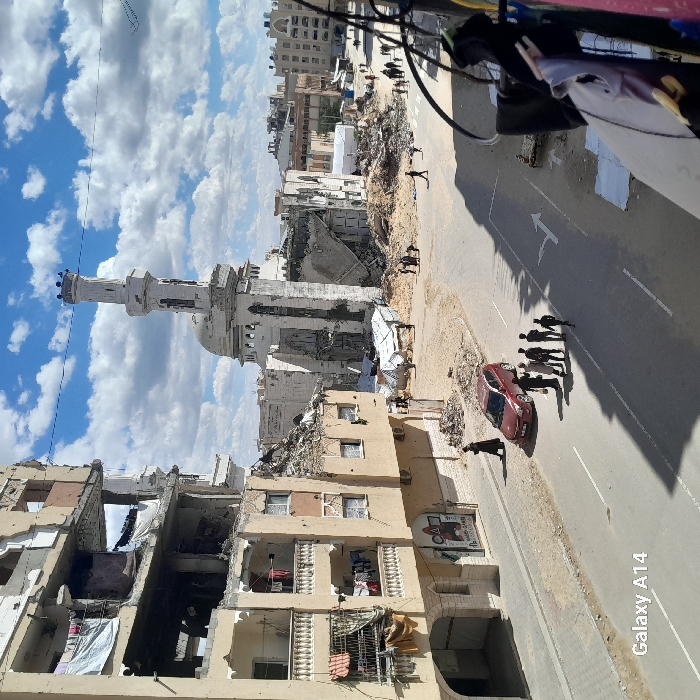 The image presents a scene of extensive urban destruction in Khan Yunis, Palestinian Territory, under a partly cloudy blue sky. In the left foreground, a multi-story building exhibits severe structural damage, with upper floors partially collapsed and interiors exposed; laundry is visible hanging on balconies. A paved road with a white directional arrow runs in front of this building. On the road, a dark red sedan is parked, and a group of approximately six individuals, predominantly adult males, are gathered nearby. A solitary figure in dark clothing walks on the sidewalk alongside the damaged building.
The central and right midground consists of a large expanse of rubble and debris, indicative of widespread building collapse. Numerous individuals are scattered across this area, some walking, others seemingly observing the damage or sifting through debris. Several temporary shelters, constructed from white tarpaulins or corrugated metal, are present within this damaged zone. Dominating the background is a severely damaged white mosque or religious structure, featuring a tall, partially intact minaret with damage to its lower sections, and a largely destroyed main building and dome. Further in the background, additional multi-story buildings, some appearing damaged, line a street where more pedestrians are visible. The scene is illuminated by direct daylight, casting clear shadows.