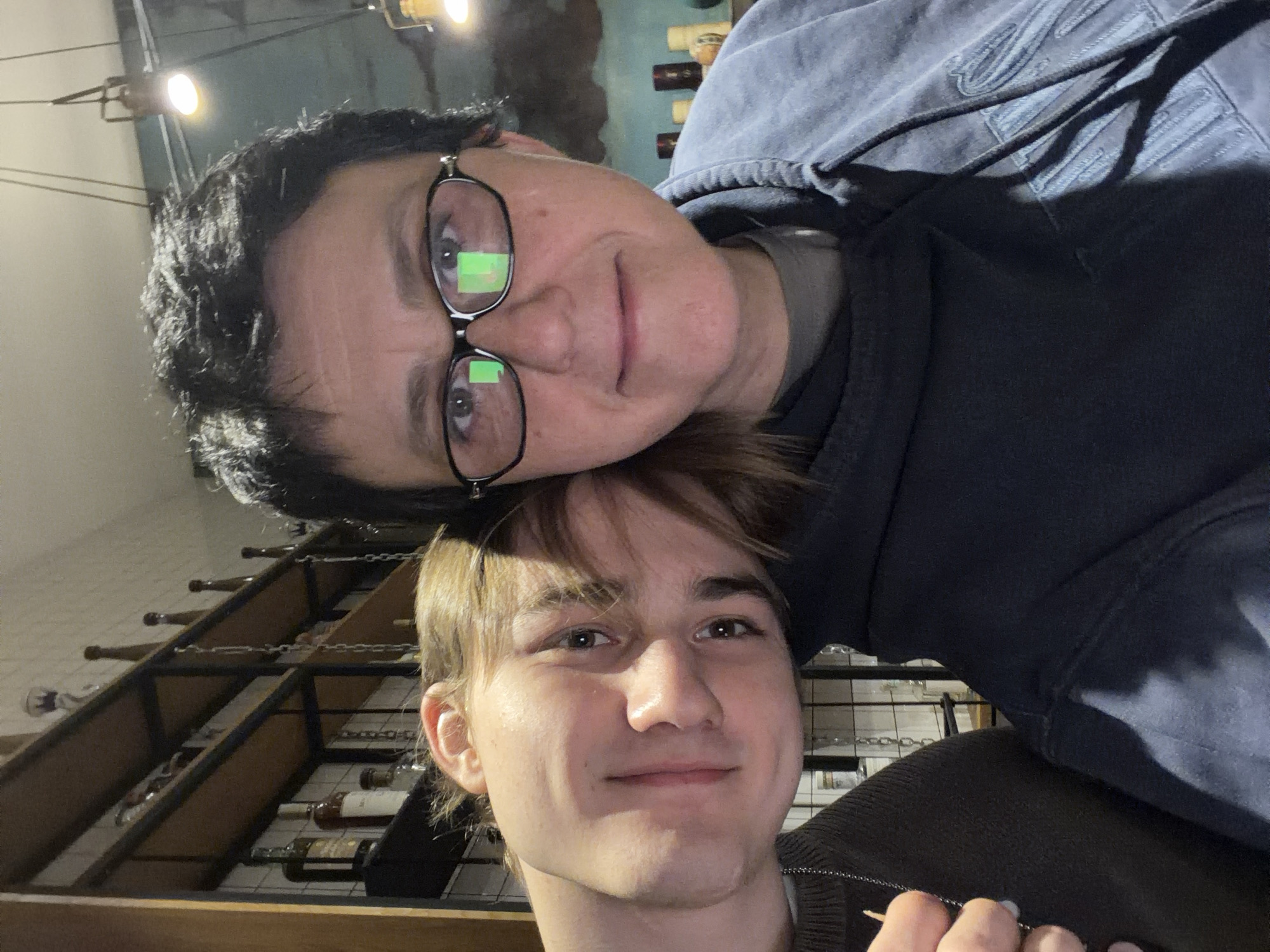 Two people are posed closely together in what appears to be a cellar setting. The lower figure is a fair-skinned individual with light brown hair styled in a side part; they are smiling slightly and face the camera. The upper figure has short, dark curly hair and wears glasses. They have a blue hooded sweatshirt on. The background features a metal wine rack filled with wine bottles. This suggests the setting could be a wine cellar or storage area associated with a restaurant, shop, or private residence. Tbilisi, Georgia, is not explicitly verifiable from the given visual data.