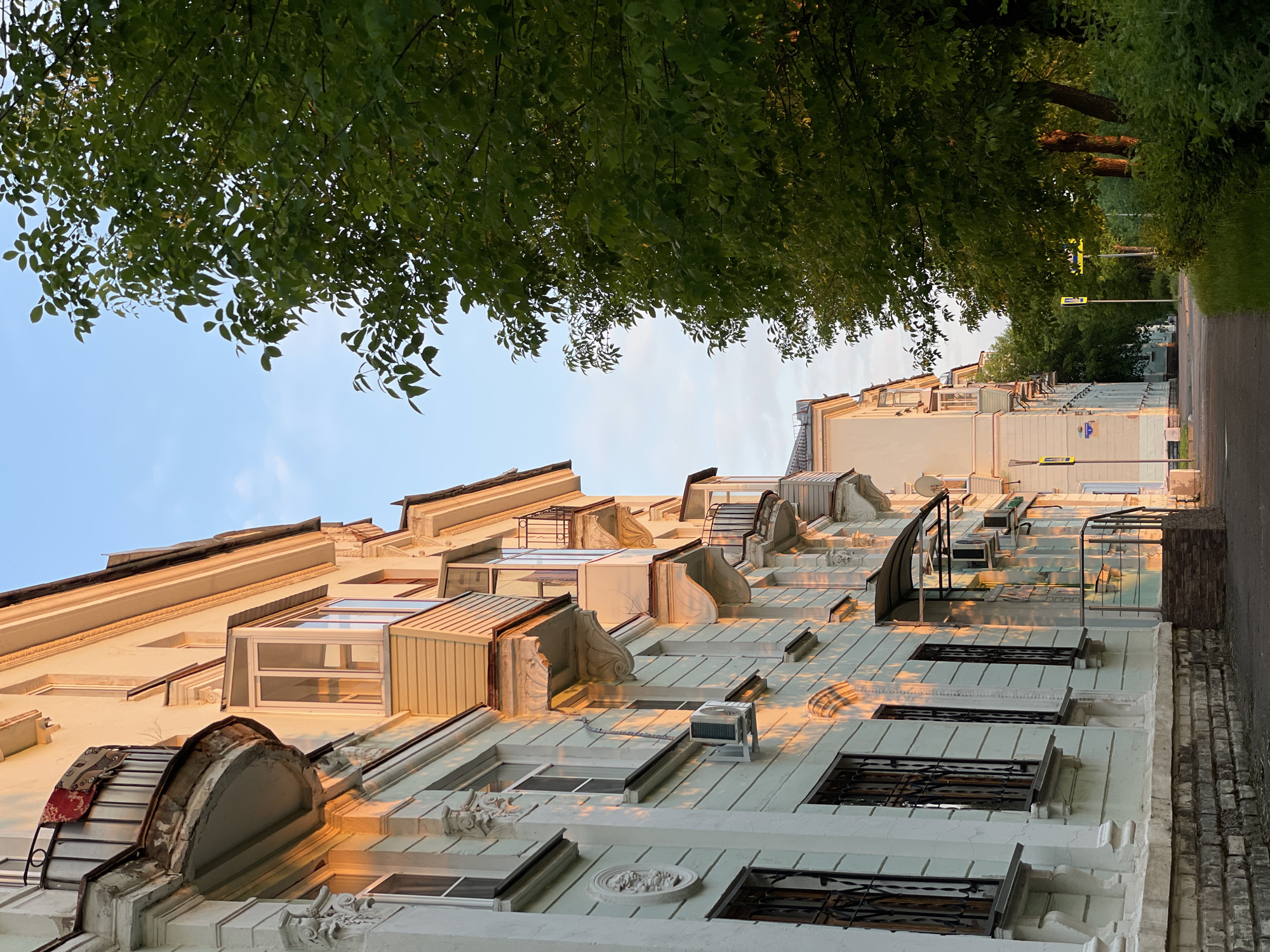 The image depicts a street scene in Zheleznogorsk, Russia. On the left, an apartment building with a pale green facade is visible, featuring decorative architectural details, windows with dark metal grates, and small balconies. The late afternoon sun casts warm, golden light on the building. To the right of the building, a sidewalk leads away from the viewer, bordered by a dense canopy of green trees. The sky above is a clear blue with a few wispy clouds. Further down the street, more apartment buildings and trees can be seen, suggesting a residential area. The overall impression is of a tranquil urban environment bathed in soft sunlight.