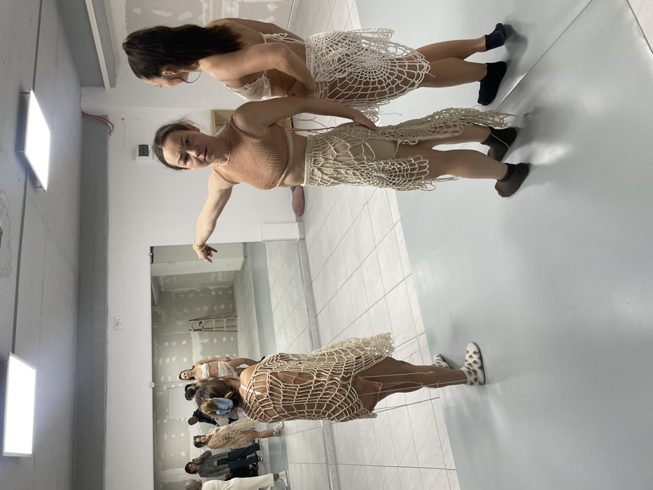 The scene depicts an indoor studio or rehearsal space located in Budva, Montenegro. The foreground features a light gray floor section, while the area visible beyond it and in the large mirror reflection on the left has light-colored tiled flooring. The left wall is dominated by this mirror, reflecting the opposite side of the room, which appears to be in an unfinished state with exposed plasterboard walls and a visible wooden stepladder. The ceiling is light gray and fitted with two rectangular LED panel lights.
Three women are prominent in the foreground and midground. In the foreground-right, one woman wears a light brown knitted crop top and a long, light beige open-knit skirt with irregular strands over an underlying light garment. She extends her right arm horizontally to the left, pointing, with a neutral facial expression. She wears dark socks. To her right, a second woman is viewed from the back-right, wearing a white open-knit bikini top and a matching skirt over a light undergarment. Her dark hair is in a high ponytail, and her head is turned slightly left, observing the first woman. She also wears dark socks. To the left in the midground, a third woman is seen from the back, wearing a large, light beige open-knit poncho or shawl over a light garment. Her hair is secured with a light blue scrunchie, and she wears white, patterned slip-on shoes with white socks.
The mirror's reflection shows multiple individuals. The third woman is reflected, facing the mirror. Another woman, reflected centrally, wears a white open-knit bikini top and skirt, smiling. Several other individuals, including men, are partially visible in the reflected background, observing the scene. A small pink cushion or object is visible on the floor against the wall in the reflected area.