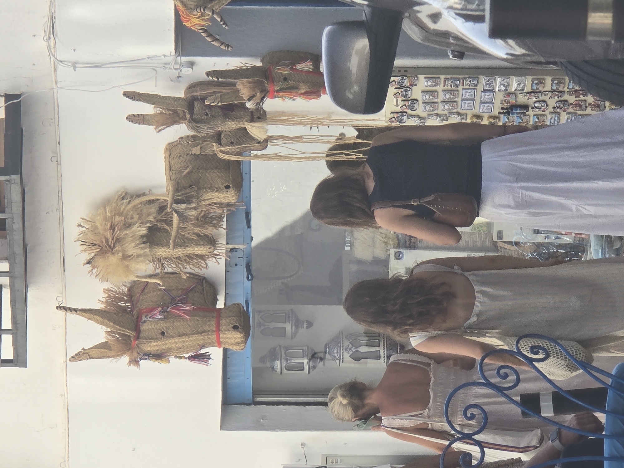 The image depicts a street scene in Mijas, Spain, characterized by its whitewashed buildings and artisanal crafts. In the foreground, three women are seen from behind, browsing. The woman on the left, with blonde hair tied back, wears a white dress and holds a phone. The central woman, with dark wavy hair, wears a light-colored dress and carries a woven bag. The third woman, also with dark hair, wears a black tank top and a long white skirt, holding a brown handbag.

Above the women, the exterior wall of a building displays several handmade straw decorations, including what appear to be stylized donkey heads with colorful tassels, reflecting the town's association with donkeys. The backdrop features typical Mijas architecture with white walls and a blue trim around an open doorway or window. Some decorative lanterns are visible inside the storefront. To the right, part of a dark-colored vehicle, possibly a car, is visible, including a side mirror and a tire. A display of small framed items, possibly souvenirs or artwork, is mounted on a wall further to the right. The overall impression is of a bustling, tourist-oriented street in a Mediterranean town.