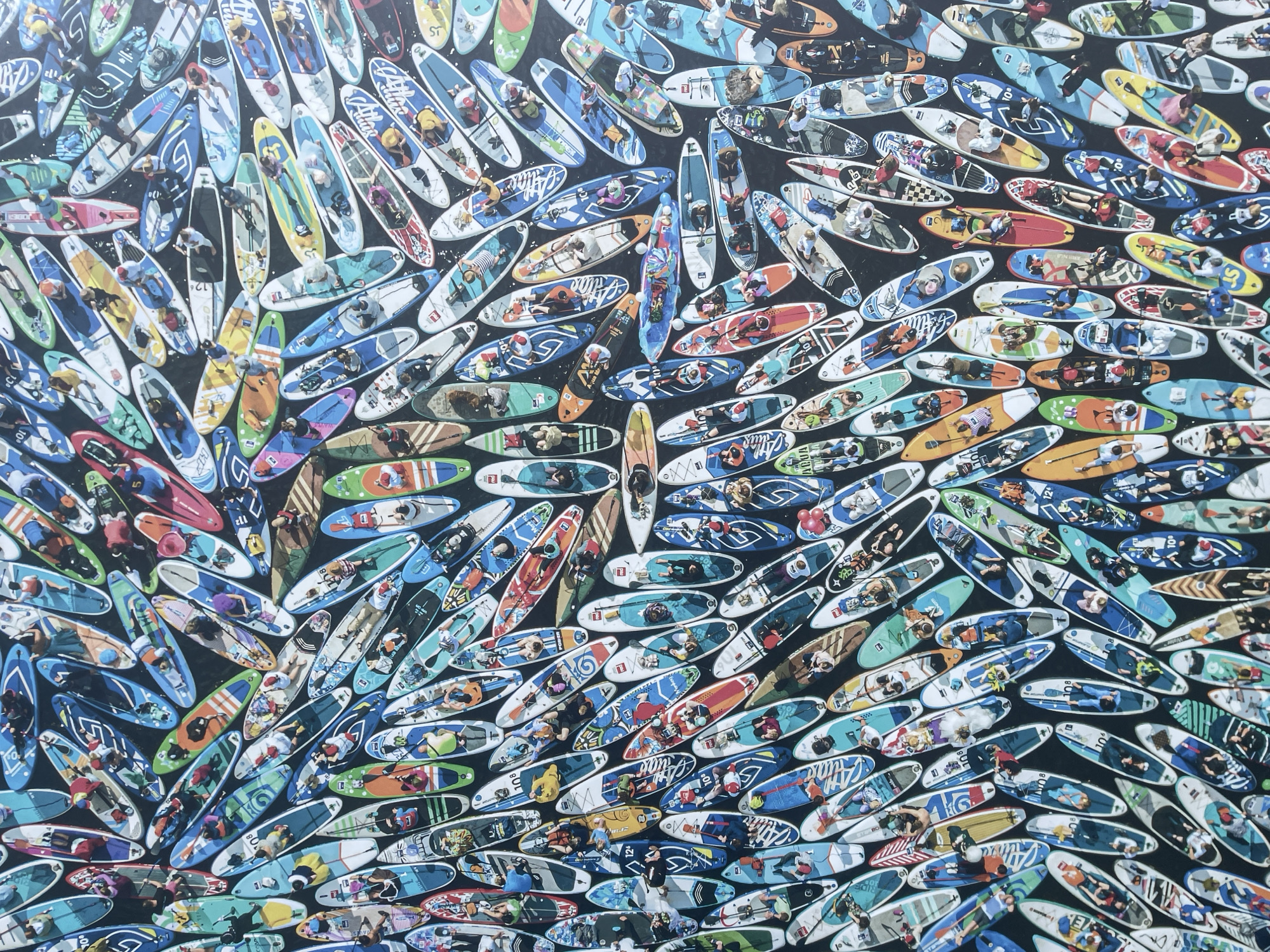 The image shows a dense cluster of colorful paddleboards floating on dark water. Each board has one or two people on it, mostly lying down or sitting. There's a variety of designs and colors on the boards, some with numbers or logos. It is difficult to determine the location or if it's in Sochi, Russia, based on the visual information.