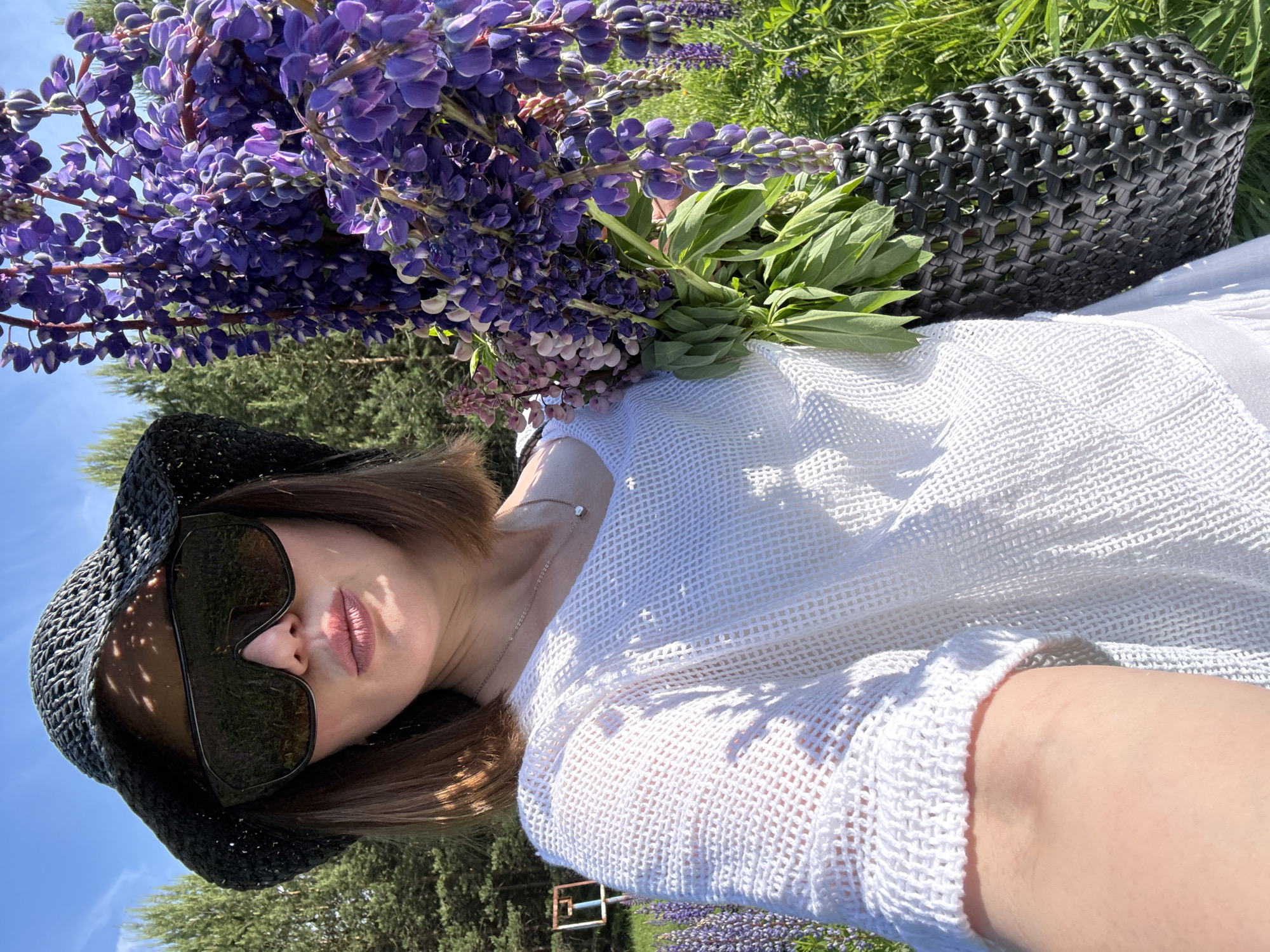 A female individual is depicted in a self-portrait, outdoors, holding a substantial bouquet of lupine flowers. She wears a black, wide-brimmed, woven hat that casts dappled shadows on her face, and oversized, dark-tinted sunglasses. Her hair is short to medium-length and brown. She is dressed in a white, loosely woven V-neck top, and a slender necklace with a small pendant is visible. The lupine flowers are predominantly purple with some pinkish-purple accents, and are held within a black, woven bag resembling a tote or basket. The background consists of a clear blue sky, numerous green coniferous trees, and a field of green grass with additional blooming lupine flowers. A light-colored, possibly metal, frame structure is partially visible in the distant left background. The scene is brightly illuminated by direct sunlight, casting distinct shadows. The setting is a natural outdoor environment, in what is specified as the city of Chaikovsky, Russia.