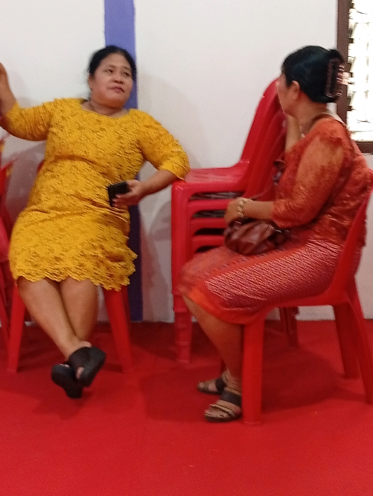 In City of Gunungsitoli, Indonesia, two women are seated on red plastic chairs, facing away from the camera. The woman on the left wears a yellow, textured, short-sleeved dress and black slip-on shoes. She holds a dark object, possibly a phone, in her right hand. The woman on the right wears an orange, patterned, long-sleeved dress and sandals with straps. She has a brown purse on her lap and her hair is pulled back with a clip. The environment appears to be indoors with a white wall in the background and a red carpeted floor.
