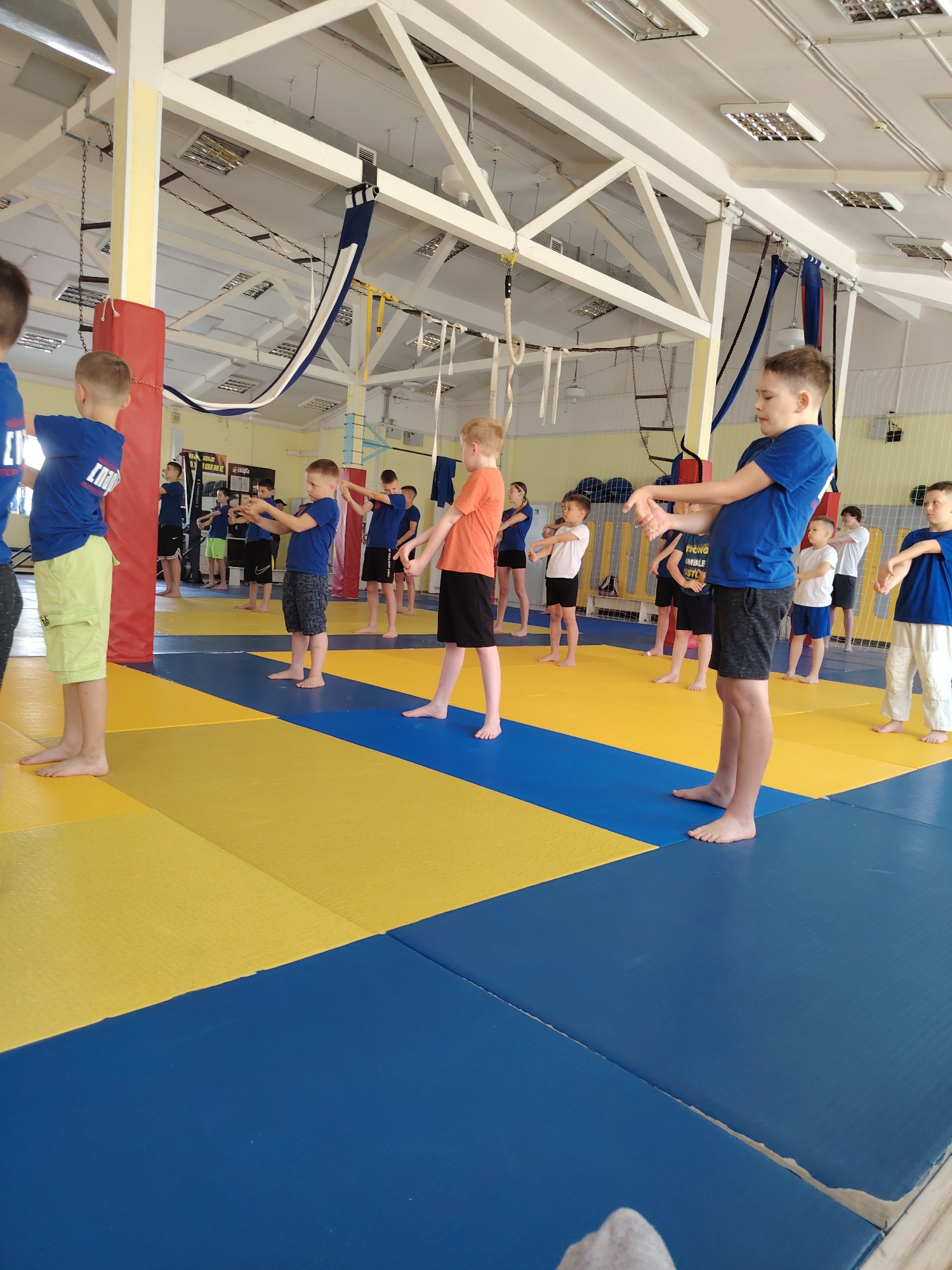 A group of children are exercising barefoot on a yellow and blue mat in a gym in Chaikovsky, Russia. The children are of varying heights and are wearing a mix of t-shirts and shorts. They are standing in rows, facing towards the right side of the image, and appear to be performing wrist stretches. The gym has high ceilings with exposed beams, and various gymnastic apparatus are suspended from the ceiling. There are columns and other equipment in the background.