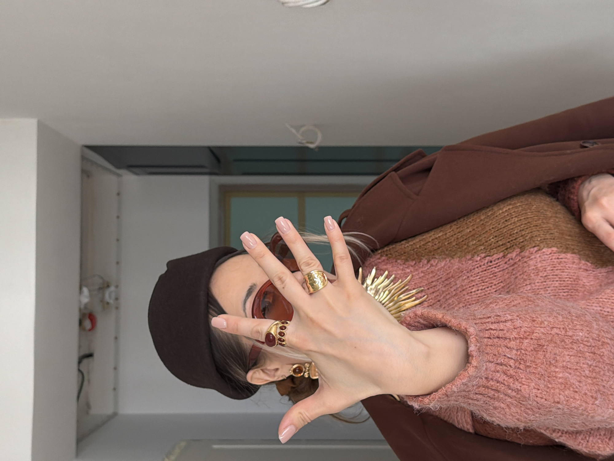 A woman wearing a brown beret, sunglasses, brown sweater, and brown jacket is indoors. She has multiple rings on her fingers. The background appears to be a residential interior, possibly an apartment. There is a white wall and ceiling. The location in the city of Krasnoyarsk, Russia, cannot be determined from this image.