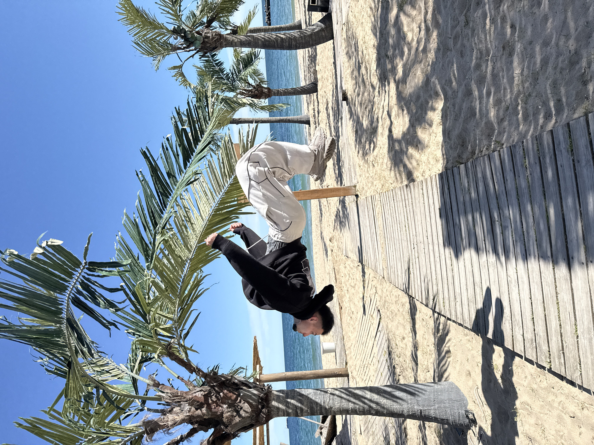 The image captures a vibrant, sun-drenched beach scene, likely in Varna, Bulgaria. The central subject is a male individual performing an acrobatic move, appearing to be an inverted handstand or balance, with his hands placed on a low wooden structure. He is wearing a black long-sleeved top or hoodie and light-colored (white or cream) baggy trousers.
The setting is a picturesque seaside, featuring a wide expanse of clear blue sky and a calm, azure sea stretching to the horizon. Several tall palm trees with lush green fronds are prominent, framing the scene and suggesting a warm climate. Some palm trees show clusters of small, green fruits, possibly coconuts. The ground consists of light-colored sand, marked with subtle ripples, and a wooden boardwalk running through the area.
The time of day appears to be mid-morning or early afternoon, characterized by bright sunlight casting distinct, long shadows across the sand and boardwalk. The weather is clear and sunny, with no clouds visible in the sky. The overall mood conveyed is one of energy and skill, set against a serene natural backdrop. No visible text is present in the media file.