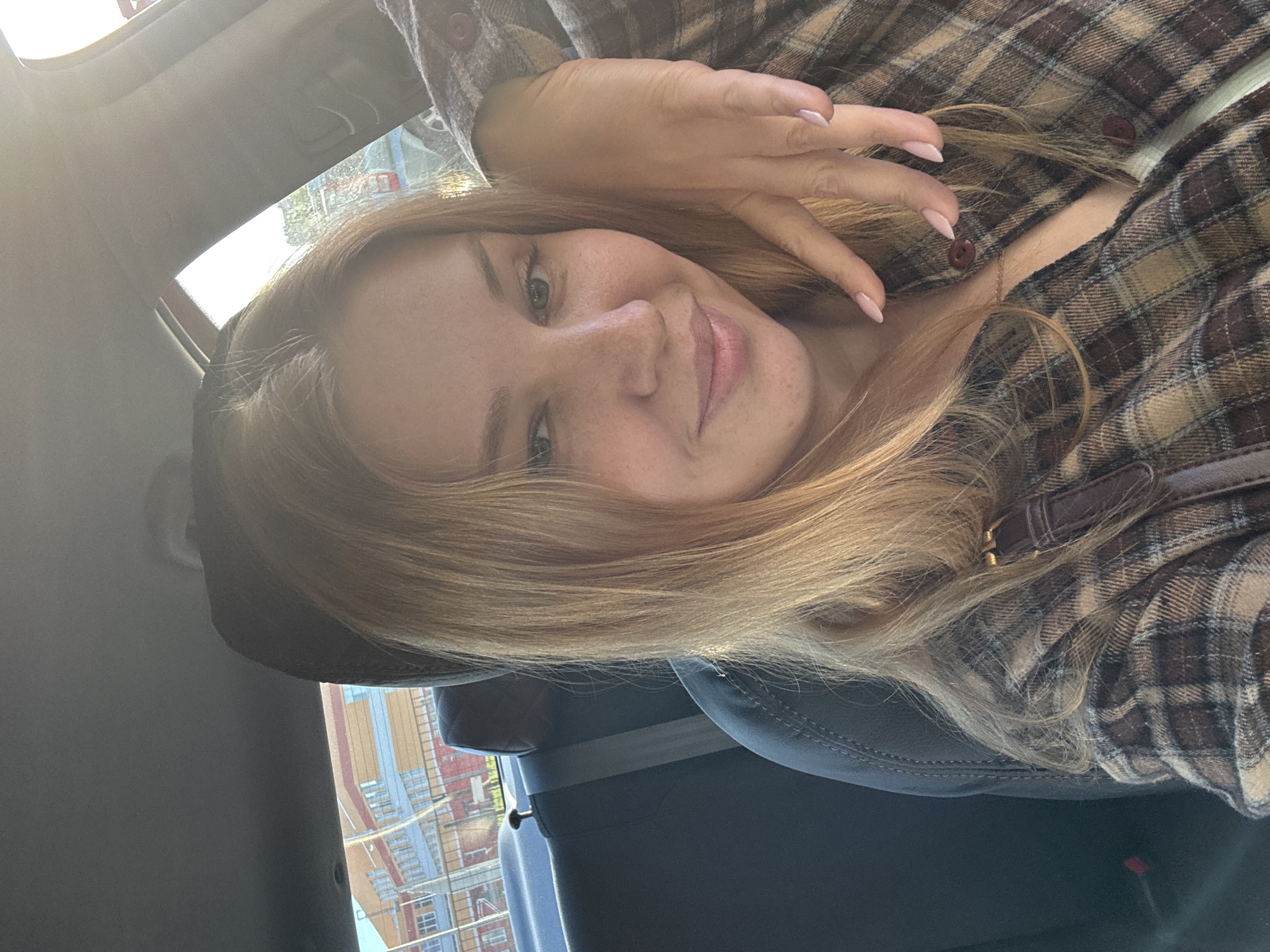 A young woman is captured in a selfie-style photograph, taken inside a vehicle, and presented rotated 90 degrees counter-clockwise. She has long, blonde or light brown hair, which frames her face. Her eyes are light-colored, possibly green or hazel, and she has visible freckles across her nose and cheeks. A soft, slight smile plays on her lips, which are a natural pink hue. Her manicured nails, painted a light pink or nude color, are visible as one hand rests near her face and hair.
She is dressed in a plaid flannel shirt, featuring a pattern of browns, tans, and creams. Dark brown buttons are visible on the shirt's front. A delicate, thin necklace chain is also visible around her neck.
The setting is the interior of a vehicle, suggested by the dark gray seat upholstery, a visible seatbelt, and the car's headliner and sun visor area. Bright sunlight streams into the vehicle from a window or sunroof, indicating it is daytime. Outside the vehicle's window, a blurry background reveals urban or suburban buildings, possibly including construction elements like scaffolding in muted tones of blue, orange, red, and beige. The overall mood conveyed is relaxed and casual. There is no visible text in the image.
