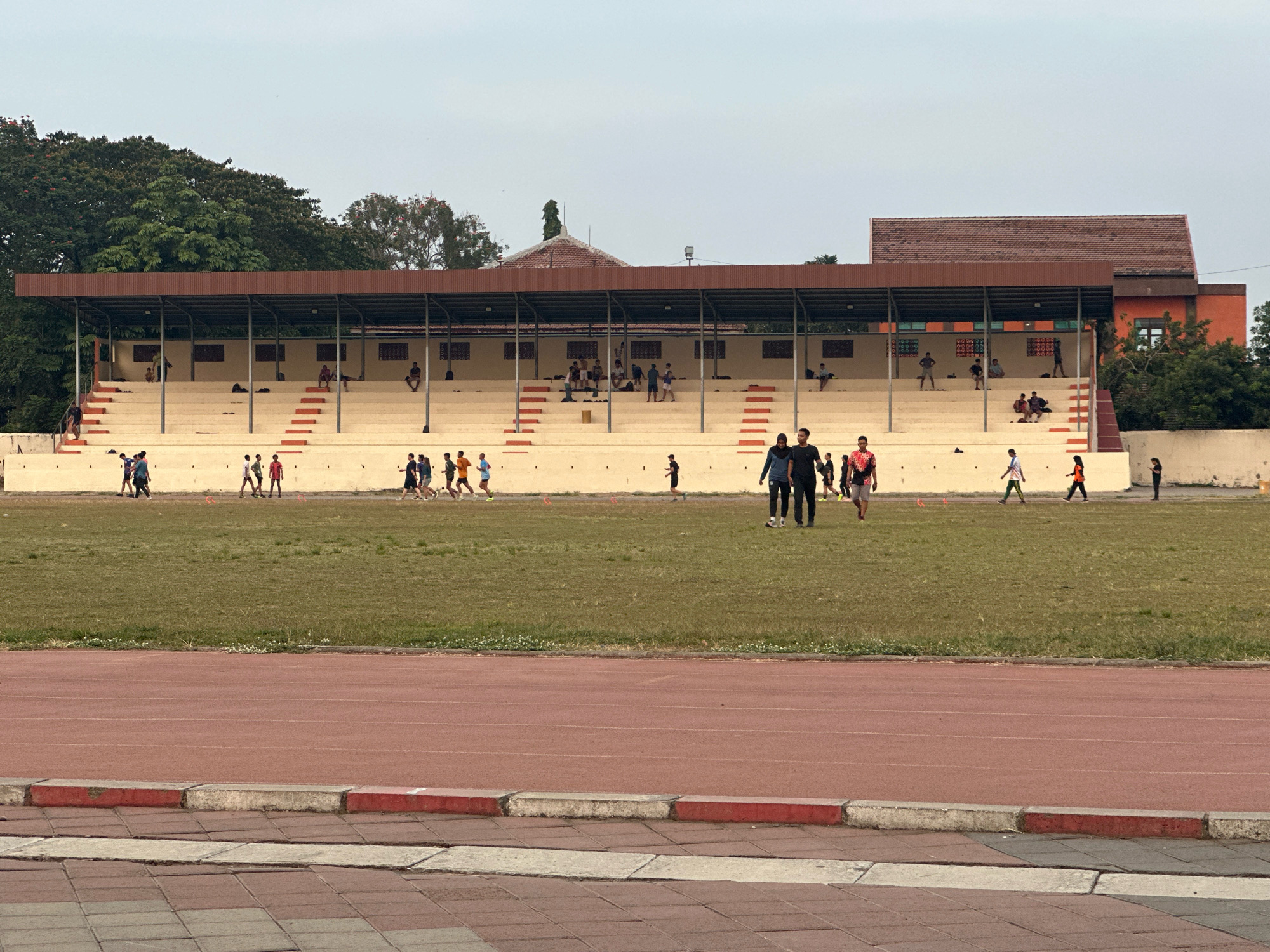 The image shows an outdoor track and field stadium in Mojokerto, Indonesia. Spectator stands with a red-brown roof are visible in the background, with people seated and standing on the bleachers. On the field, several individuals are engaged in various activities, including running and walking. Some people are gathered in small groups. The track itself is reddish-brown, and the field is covered in green grass. Trees and buildings can be seen behind the stadium. The sky is overcast.