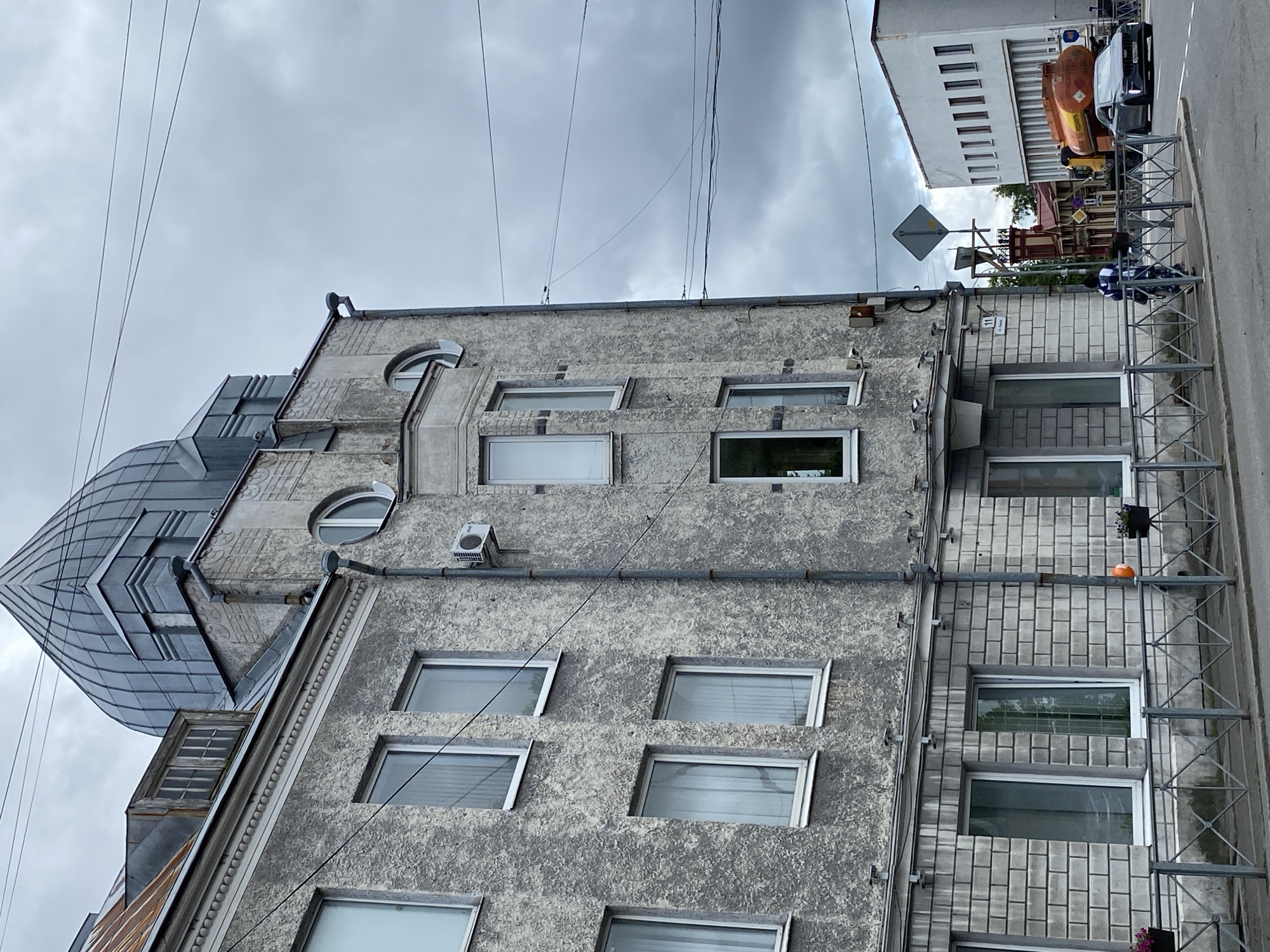 The image depicts a street scene in Sortavala, Russia, under an overcast sky. A multi-story building with a textured grey facade and a distinctive grey domed roof dominates the left side of the frame. The building features numerous windows, some rectangular and others arched. Below the main structure, the lower section is clad in light grey brickwork. A metal fence runs along the sidewalk in front of the building.
On the right side of the image, a white building with a row of rectangular windows is visible. Between the two buildings, an orange tanker truck and a dark sedan are on the road. A person wearing a backpack walks along the sidewalk towards the road. Various electrical wires crisscross the sky. The overall impression is one of an urban environment with mixed architectural styles.