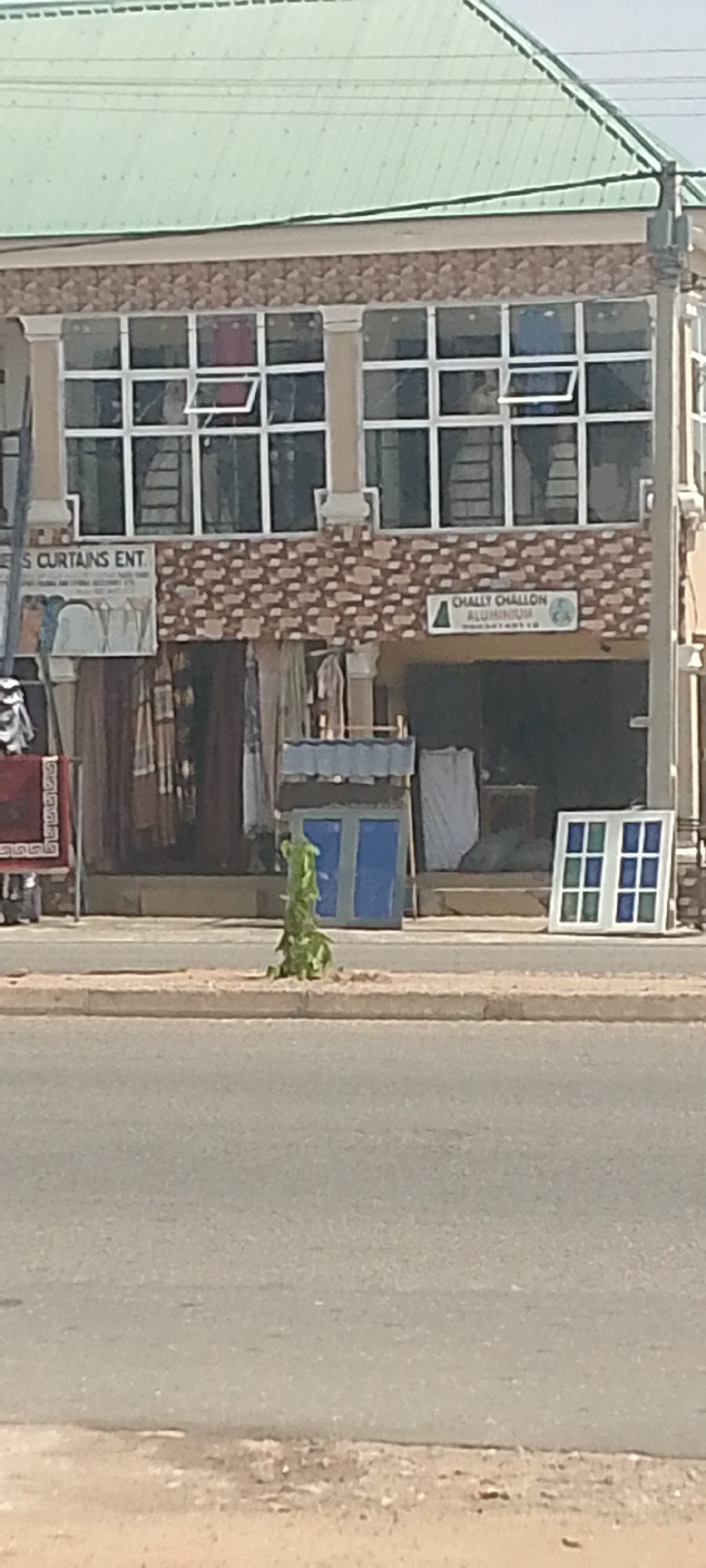The media file shows a street scene in Jalingo, Nigeria. The main subject is a two-story building with a green metal roof. The upper floor has large windows with silver frames, some of which are open. The facade of the building is decorated with a patterned brickwork.
On the ground floor, there are several businesses. One sign reads "S CURTAINS ENT". Another sign, triangular in shape, reads "CHALLY CHALLON ALUMINIUM". Beneath these signs, various items are displayed outside the shops, including what appear to be rolled-up carpets or fabrics, and some clothing or textiles hanging. There are also what look like window frames and panes displayed in front of the shops, including one with blue panes and another with green and blue panes.
In front of the building, there is a sidewalk and a road. A small, leafy green plant is growing on a patch of sandy ground between the sidewalk and the road. The overall impression is of a commercial street during daylight. The sky is visible at the top of the image, and the weather appears to be clear. The scene is likely from a daytime setting due to the lighting.