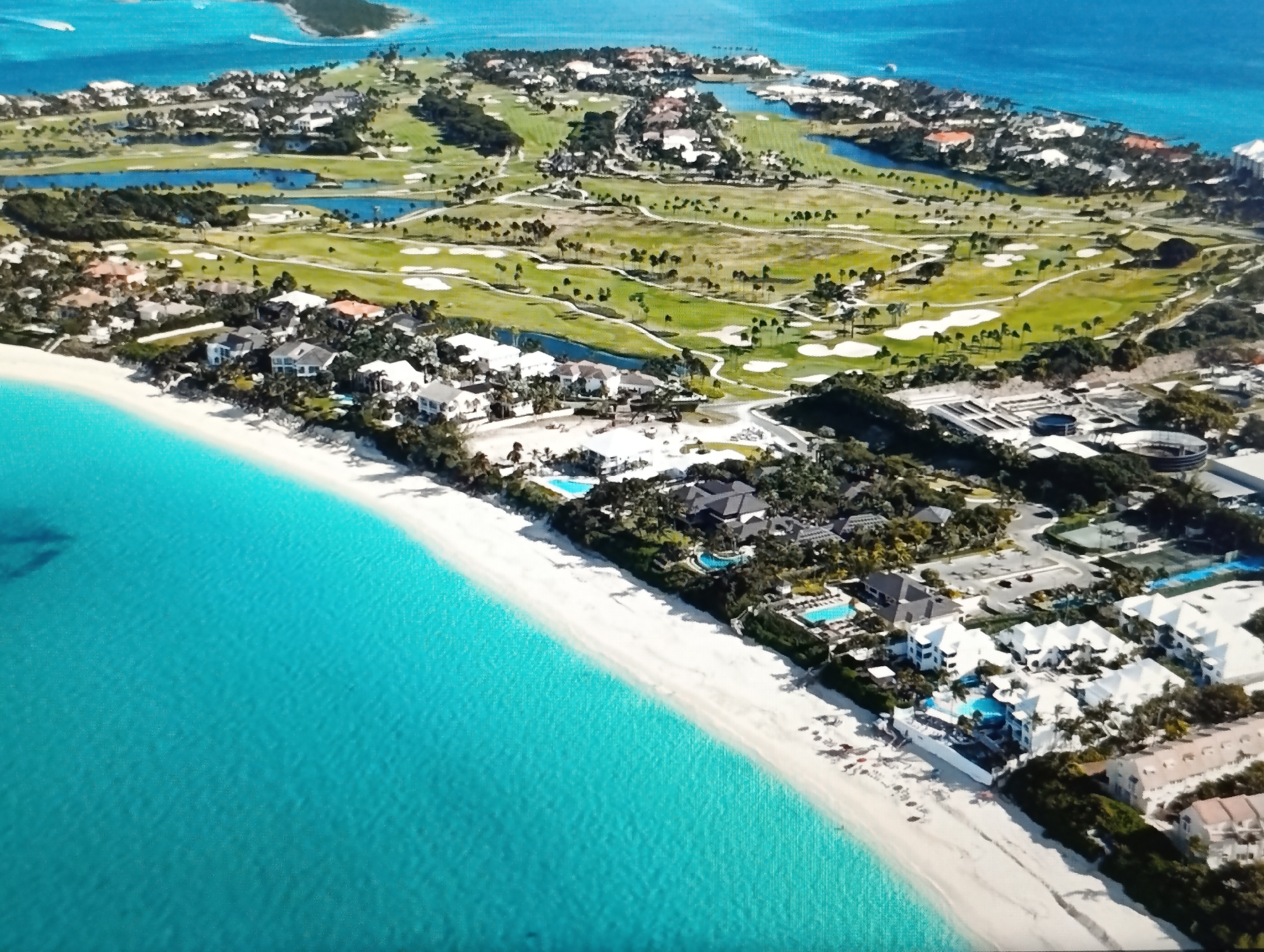The image is an aerial view of a coastal resort area featuring a white sand beach, turquoise water, and a golf course with several ponds. Numerous buildings, including villas and multi-story structures, are scattered throughout the property. Some buildings have swimming pools. A golf course with manicured fairways and sand traps is visible inland, along with several water features. The overall scene depicts a luxurious vacation destination. The image does not show any elements related to the city of Dubăsari town council, Moldova.
